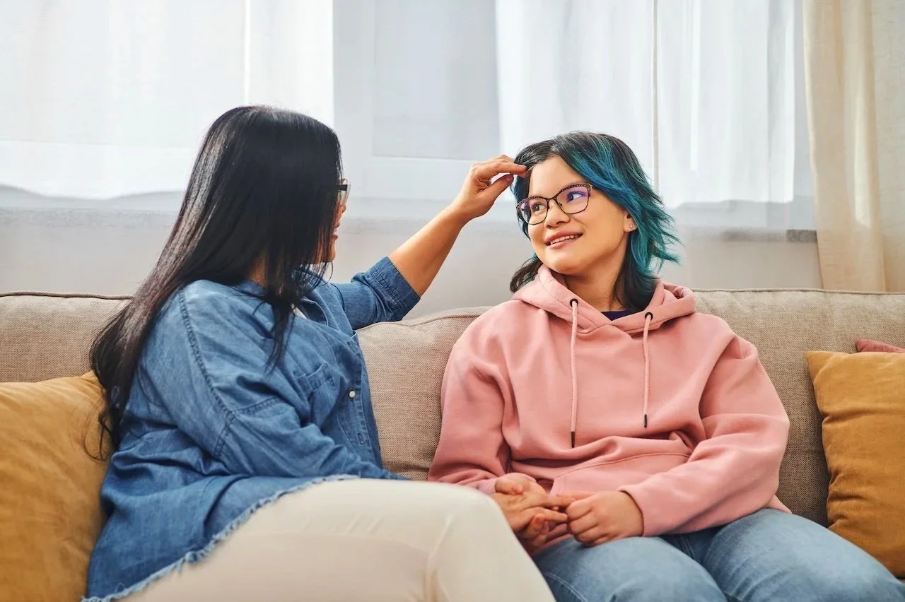 Two women sitting on a beige sofa having a conversation, with one woman touching the other's head. The woman on the right has blue and black hair, glasses, and is wearing a pink hoodie. The woman on the left has long black hair, glasses, and is wearing a blue denim jacket.