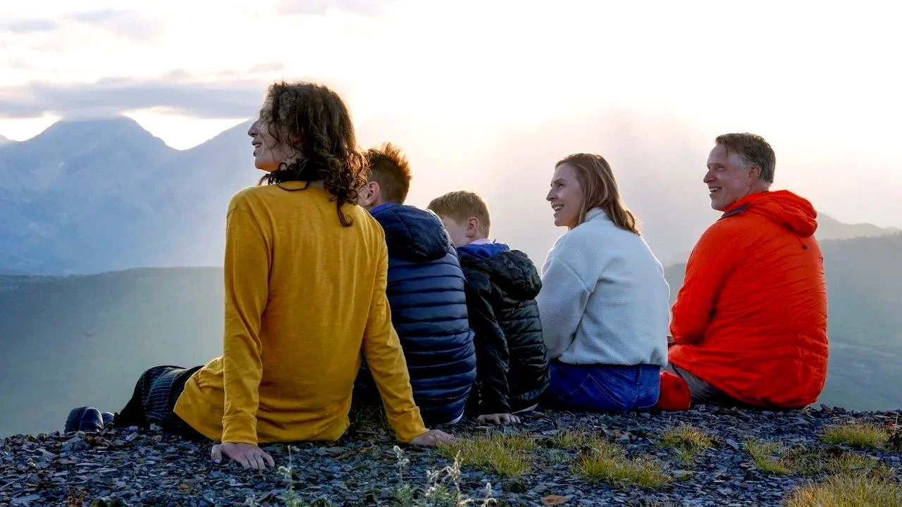 Family of six sitting outdoors on rocky ground, enjoying a mountain view at sunset, with mountains in the background.