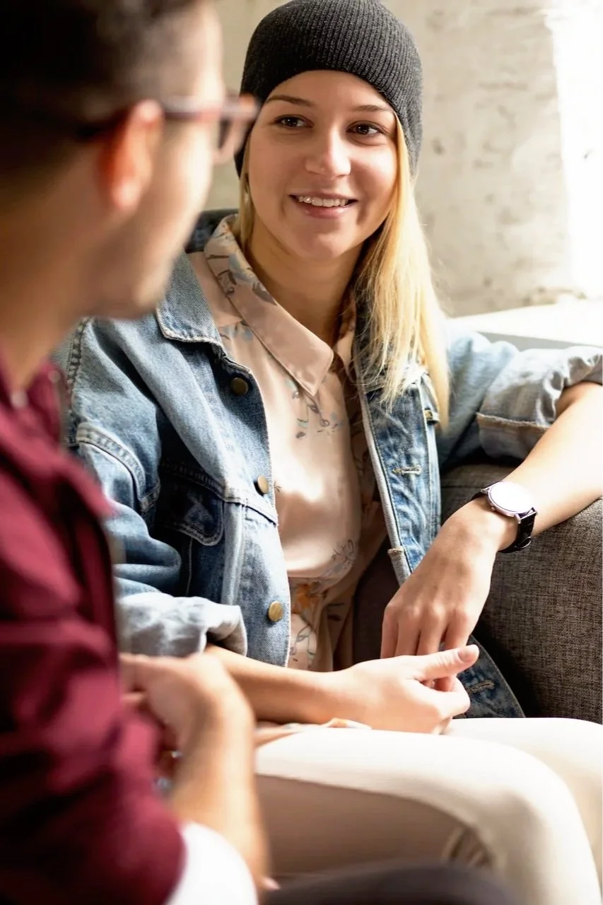 A young woman with long blonde hair, wearing a gray beanie, a denim jacket, and a floral shirt, smiling and engaging with someone in a casual indoor setting.