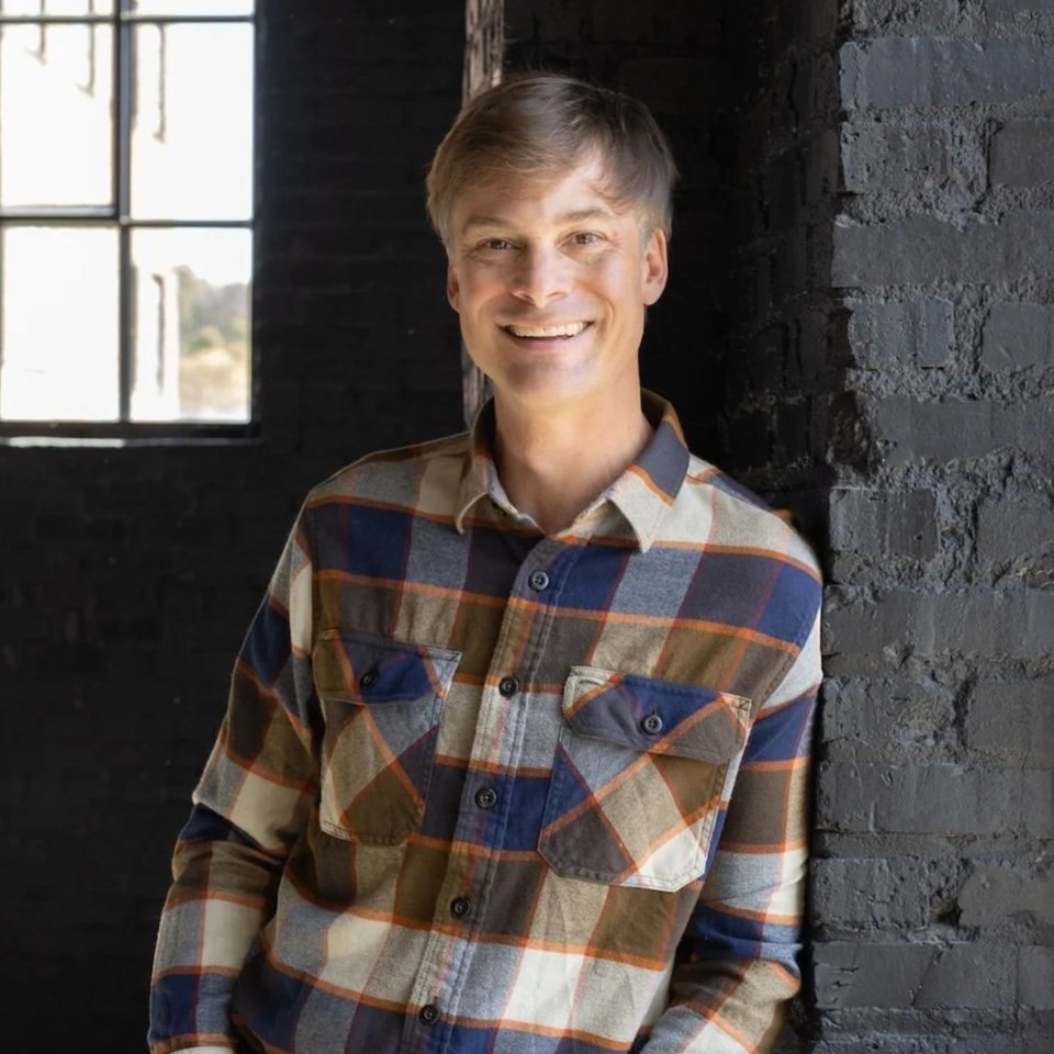Smiling man with blonde hair wearing plaid shirt leaning against a dark brick wall near a window.