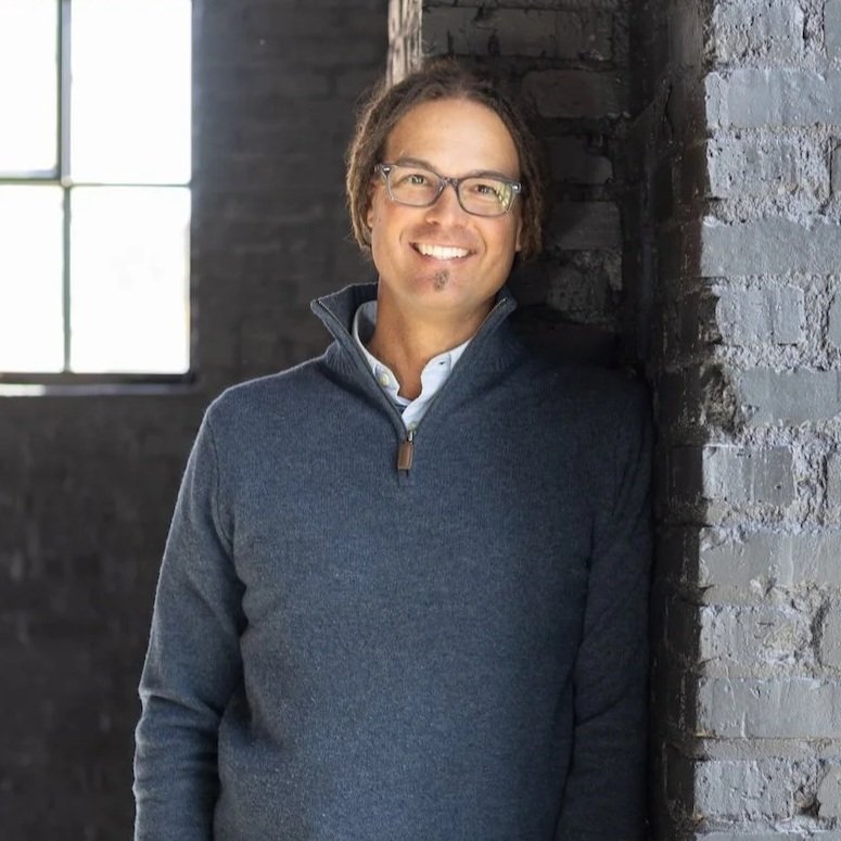 A smiling man with glasses and dreadlocks leaning against a brick wall in a room with a window.