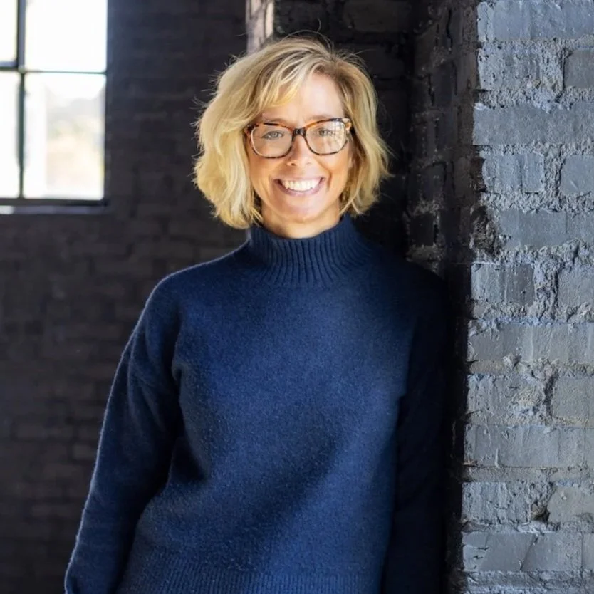 A woman with blonde hair, glasses, and a navy blue sweater smiling while leaning against a dark brick wall.