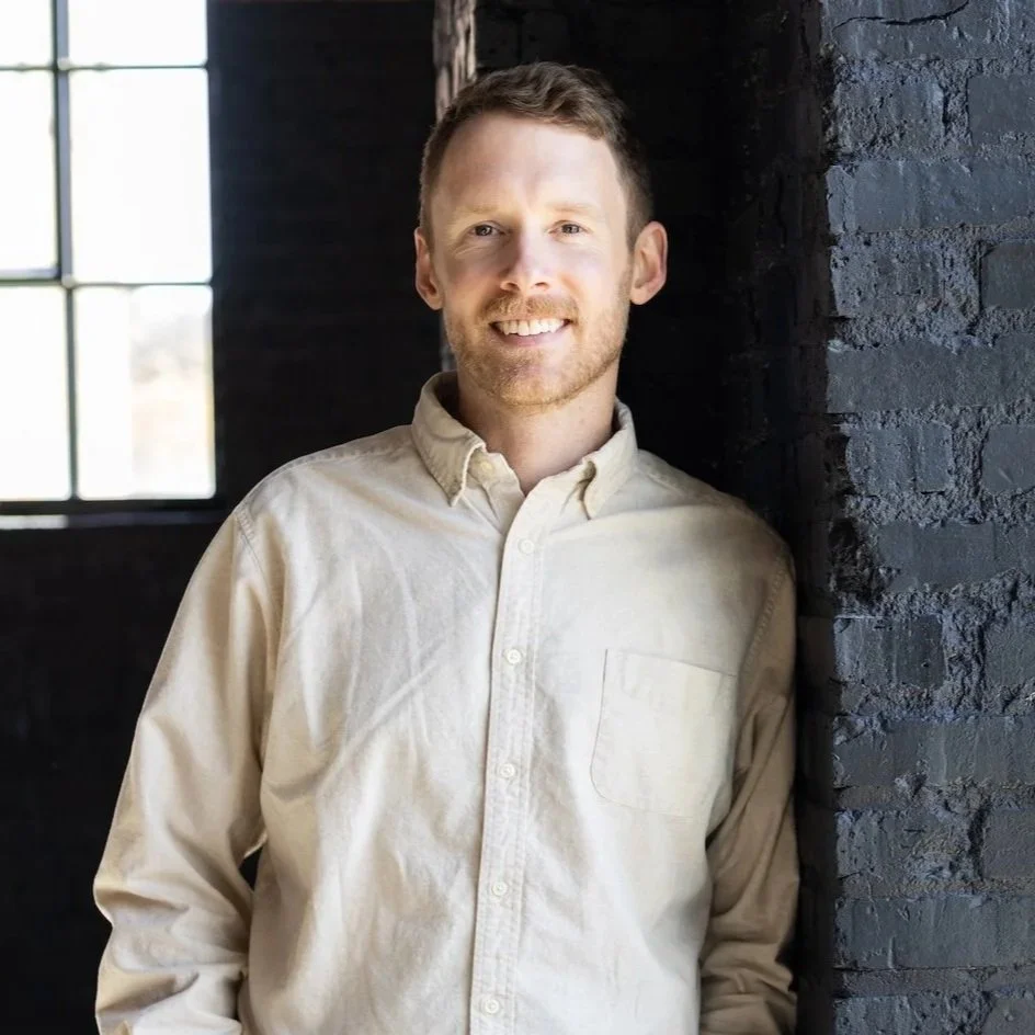 A smiling man with short brown hair and a beard, wearing a beige button-up shirt, standing beside a black brick wall inside a building with large windows.