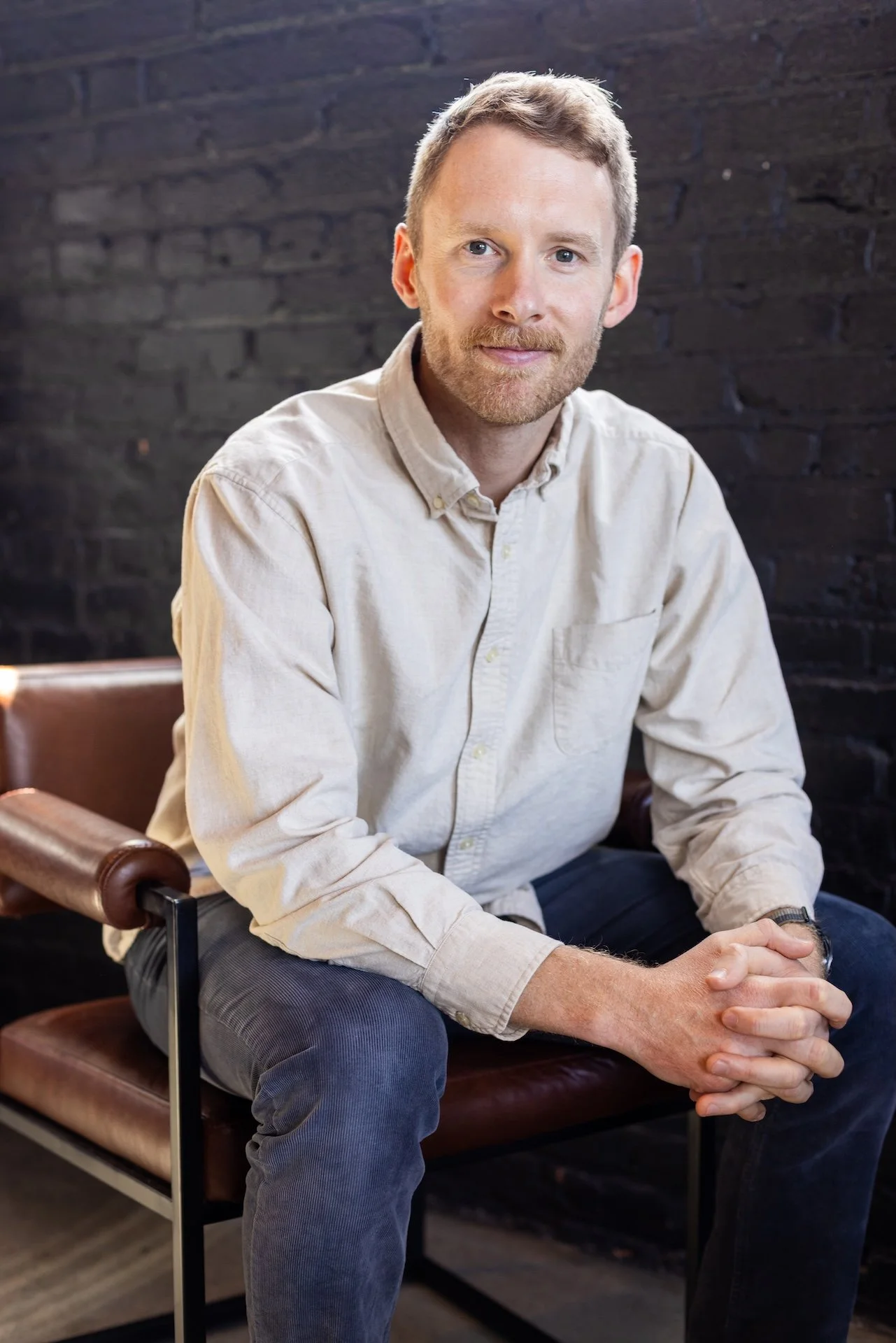 A man with light hair, beard, wearing a beige shirt and dark pants, sitting on a brown leather bench with his hands clasped, in front of a dark brick wall.