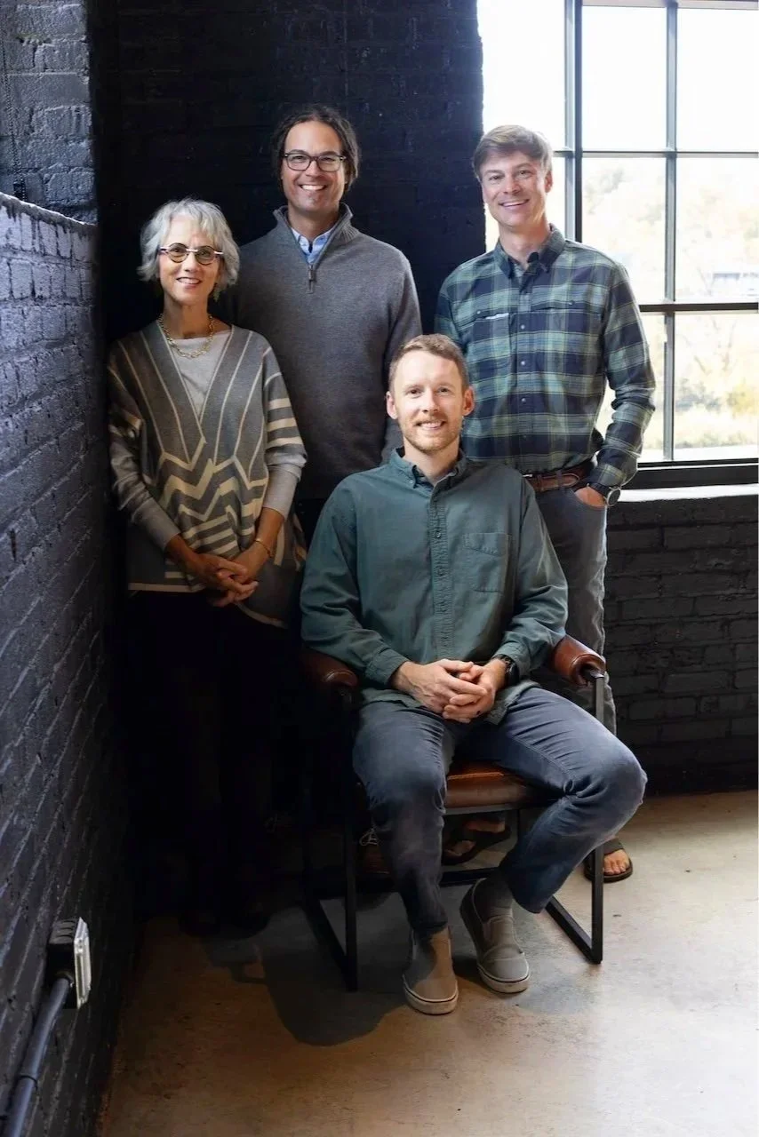 A group of four professionally dressed people posing for a photo indoors by a brick wall and large window, with one seated man and three standing individuals, smiling at the camera.