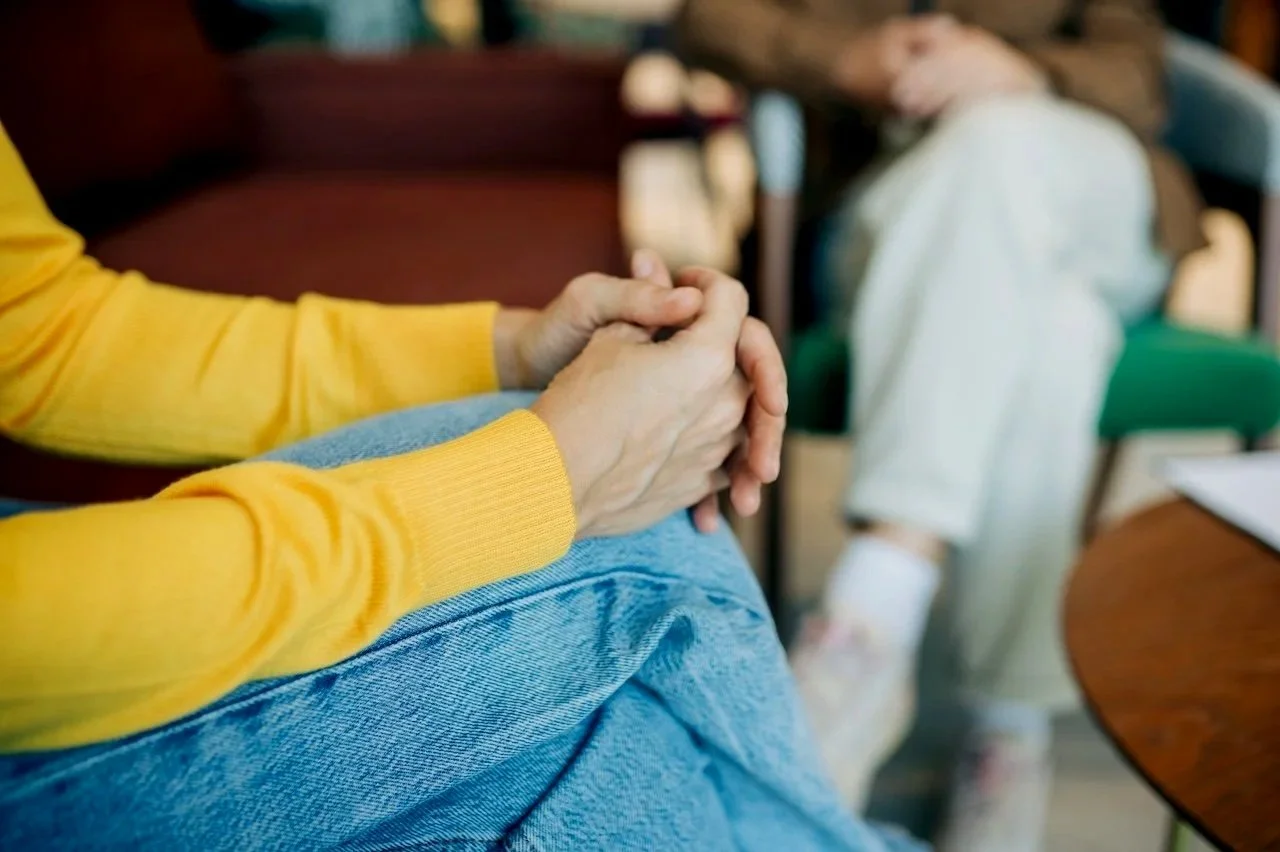 A person wearing a yellow sweater has their hands clasped together resting on their knee. In the background, a young girl with a bow in her hair is sitting on a chair, dressed in a white coat, in a cozy indoor setting.
