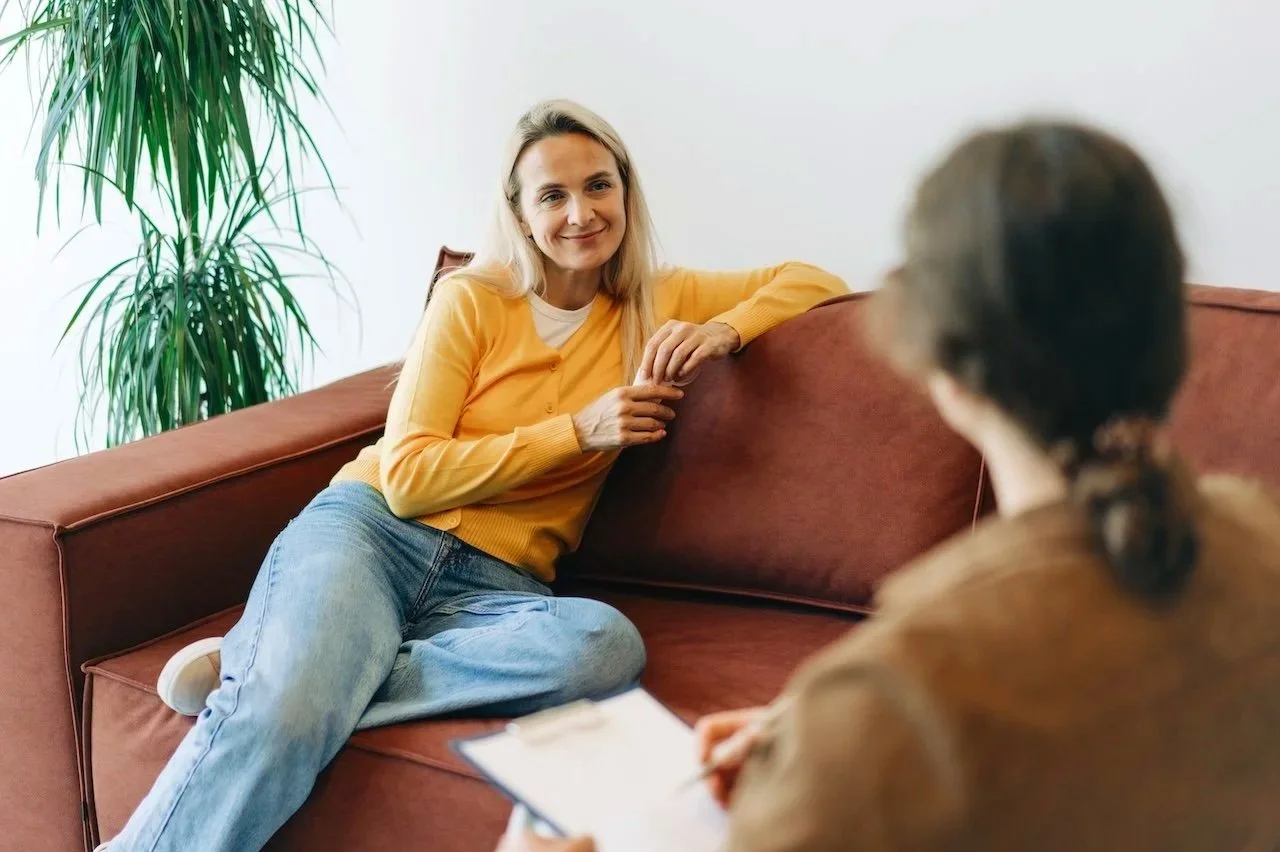 A woman in a yellow sweater sitting on a brown couch, smiling and talking to a therapist who is taking notes.