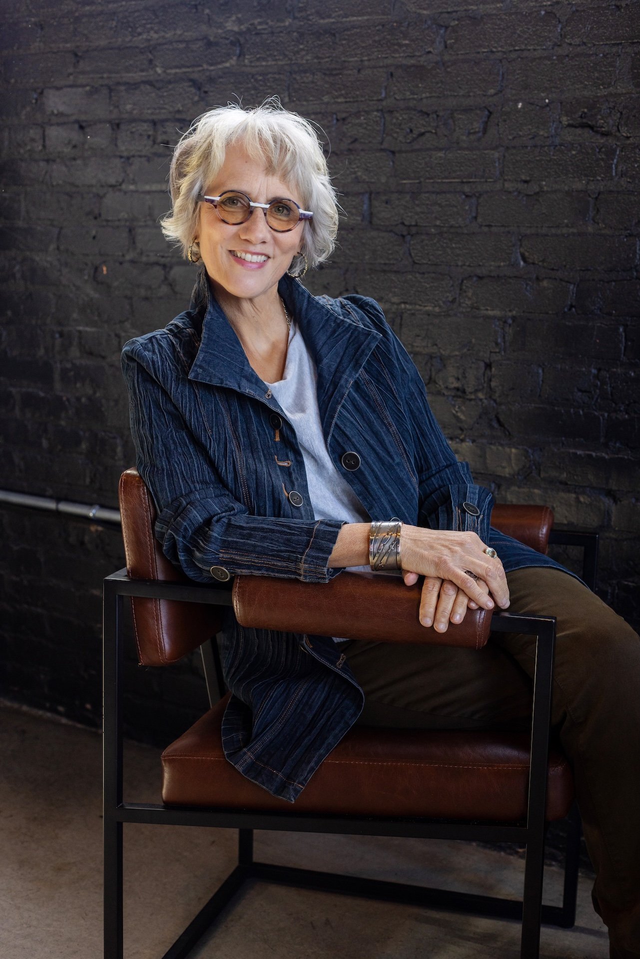 A smiling elderly woman with short gray hair and glasses sitting in a modern chair with leather armrest against a black brick wall.