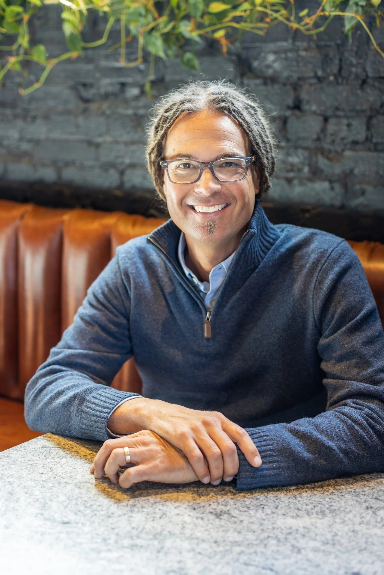 A man with glasses and dreadlocks smiling while sitting at a table in a restaurant or cafe with a dark brick wall and greenery in the background.