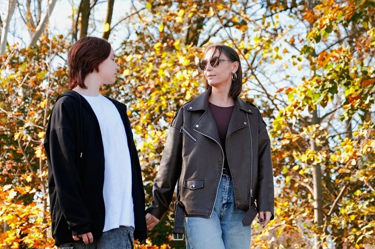A woman wearing sunglasses and a black leather jacket holding hands with a young person in a black hoodie and white shirt outdoors during fall, with trees and autumn leaves in the background.