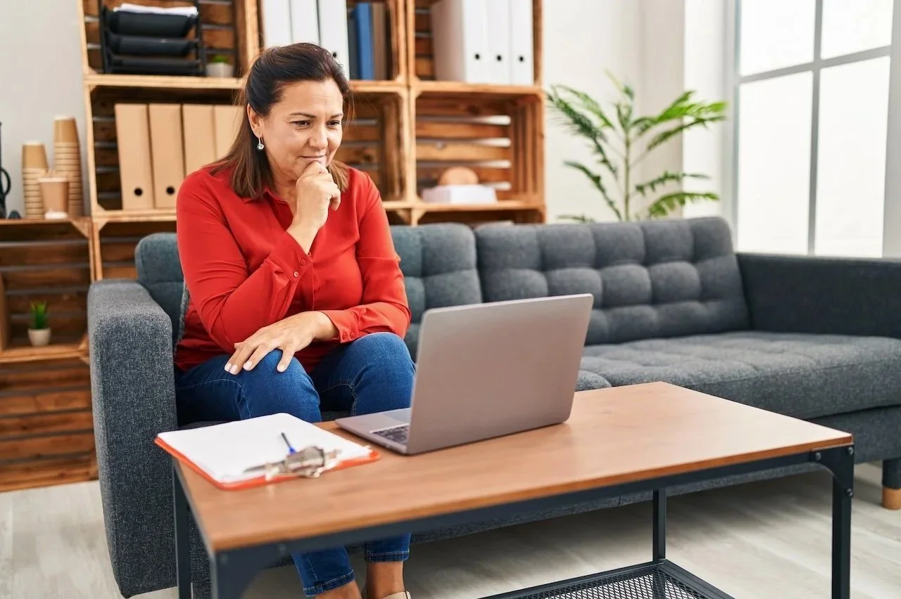 A woman in a red blouse and blue jeans sitting on a gray sofa, looking at a laptop on a wooden coffee table, with a thoughtful expression, in a modern home office or living room.