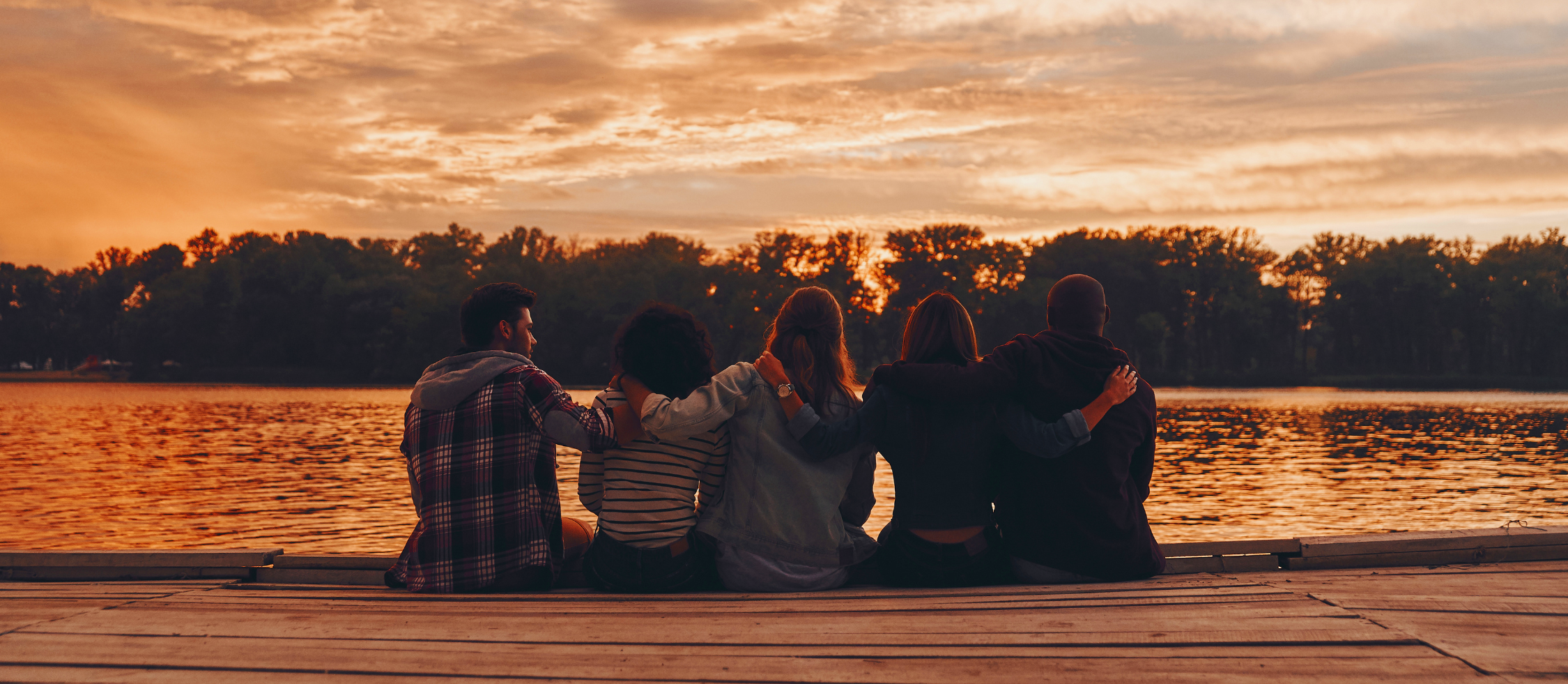 Five friends sitting on a wooden dock by a lake during sunset, with trees and a colorful sky in the background.