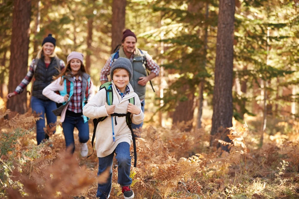 A group of four people hiking through a forest with autumn foliage. A boy in front is smiling and wearing a knit hat, fleece jacket, and backpack, leading the group. Behind him are three others, including a man and two girls, also carrying backpacks, enjoying the outdoor walk.