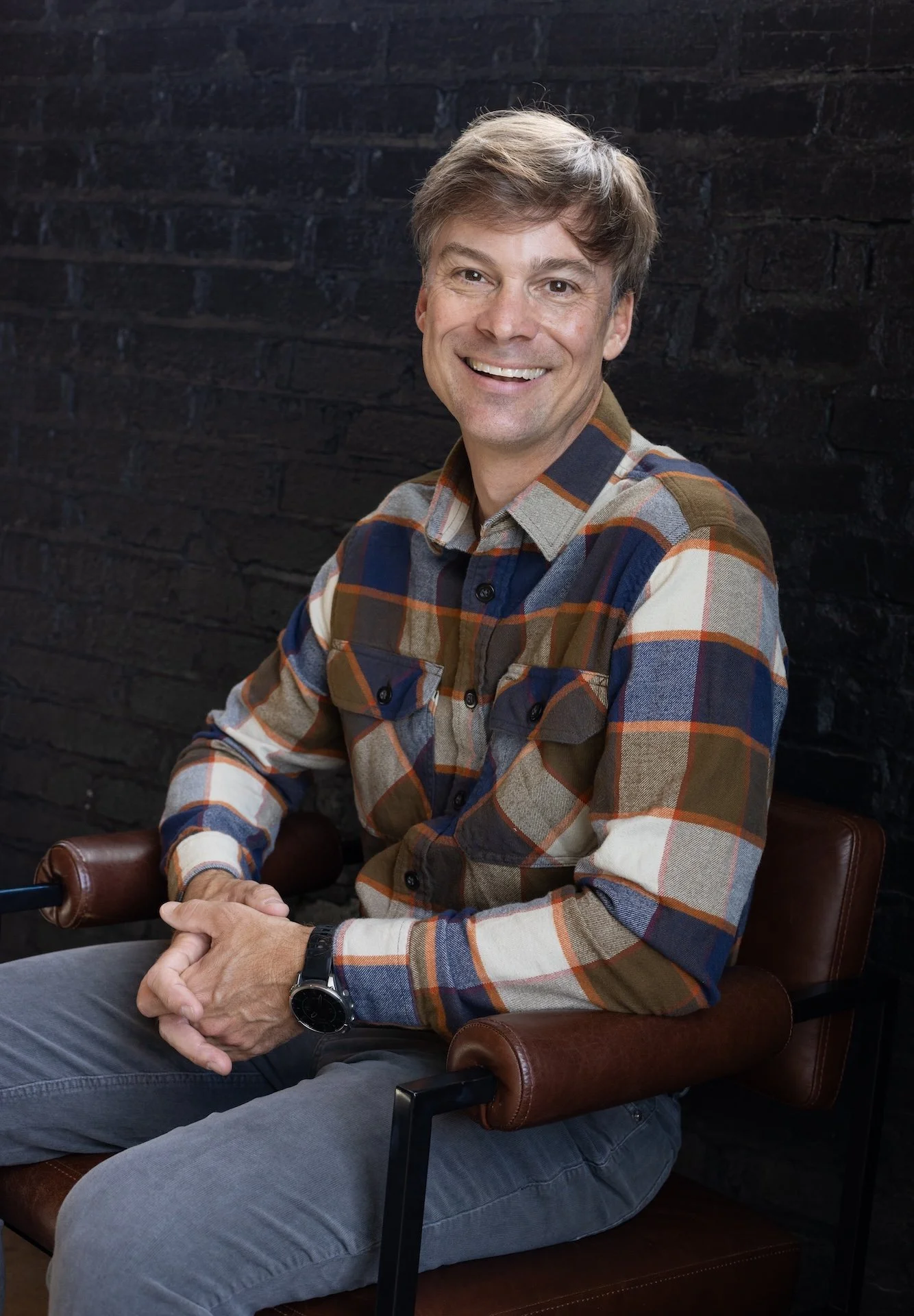 A smiling man with light brown hair, wearing a plaid shirt in shades of brown, blue, and white, seated on a brown leather chair against a black brick wall.