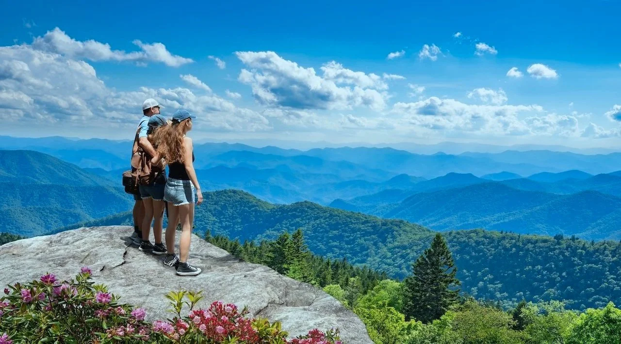 Four hikers standing on a rocky ledge overlooking a mountain range with green forests and a blue sky with scattered clouds.