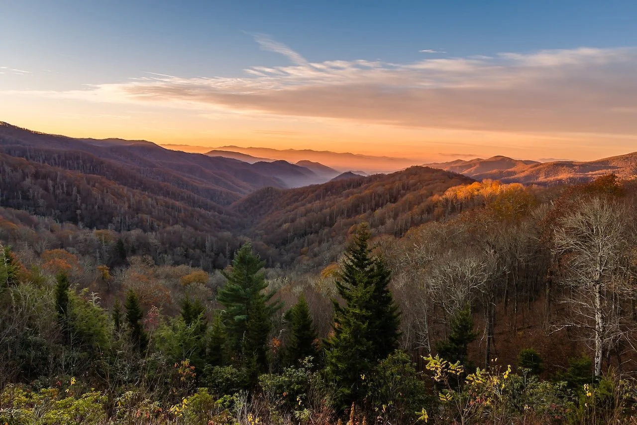 A scenic view of a mountain range during sunset with layers of forested hills, some trees with autumn foliage, and a partly cloudy sky.