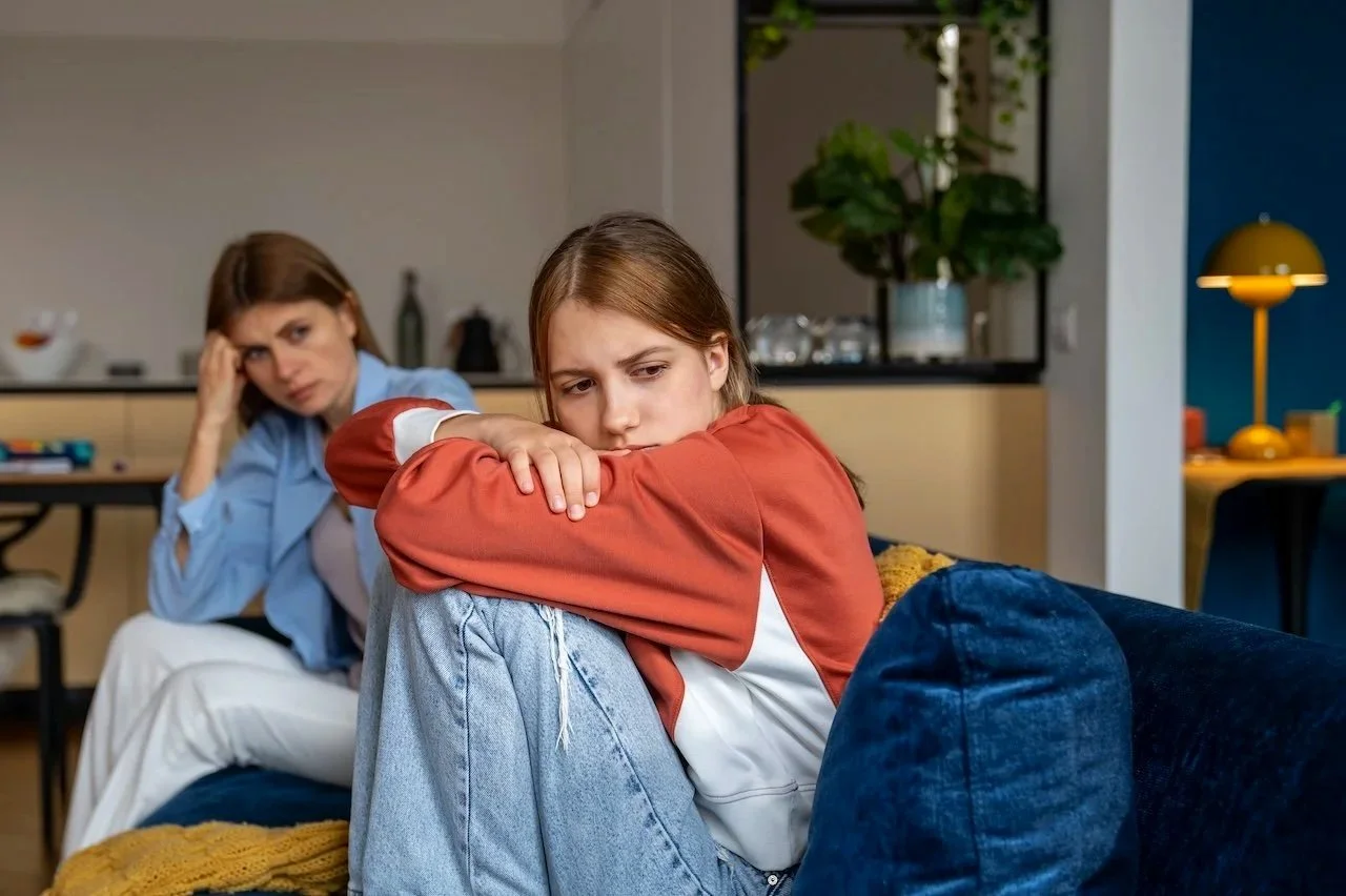 A young girl sitting on a blue couch looking distressed, with her arms resting on her knees, and a woman sitting in the background looking worried and stressed, in a modern living room.