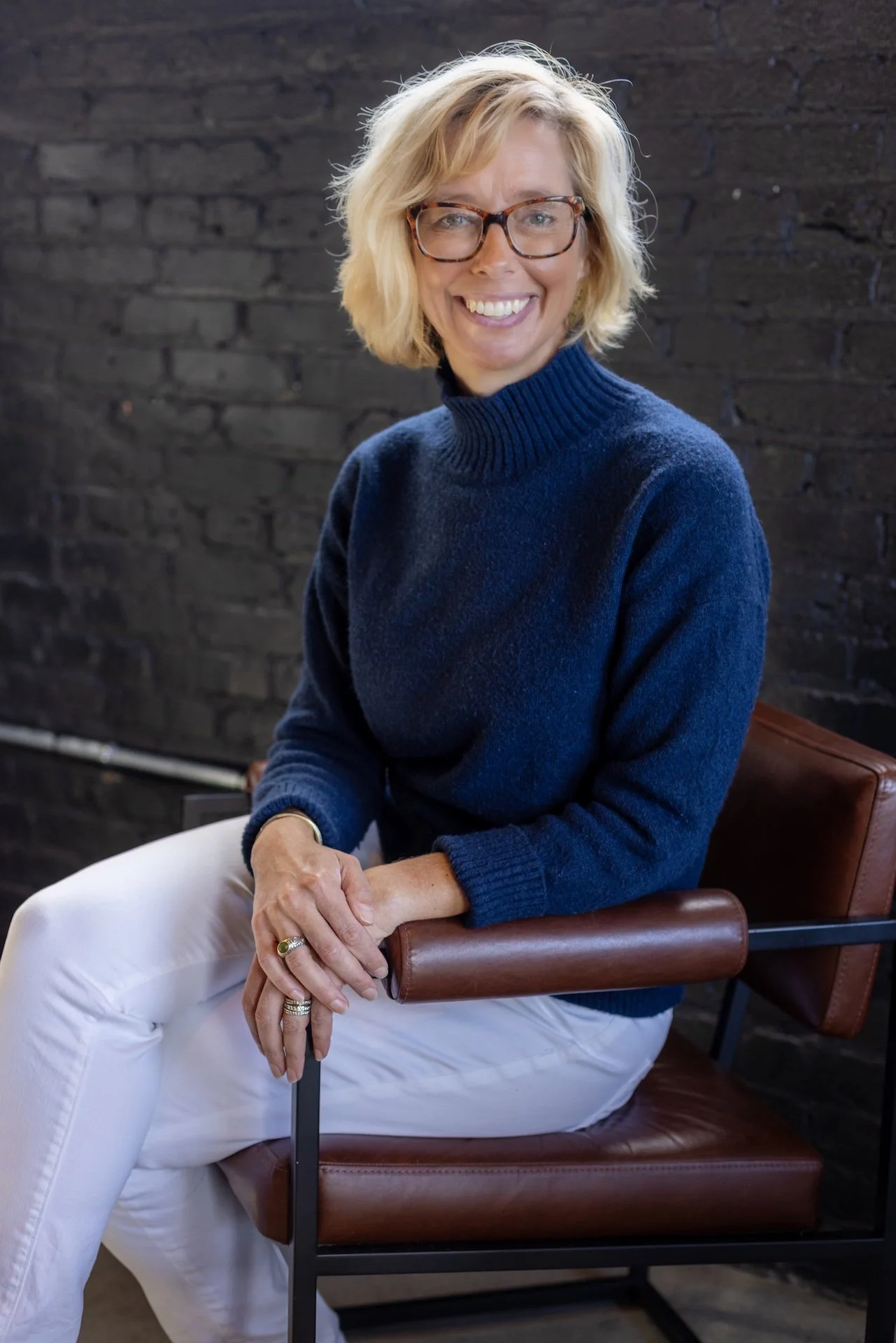 A smiling woman with blonde hair and glasses, wearing a navy blue turtleneck sweater and white pants, seated on a brown leather chair against a dark brick wall.