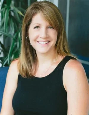 A smiling woman with shoulder-length light brown hair wearing a black sleeveless top, standing in front of a green plant and glass wall.