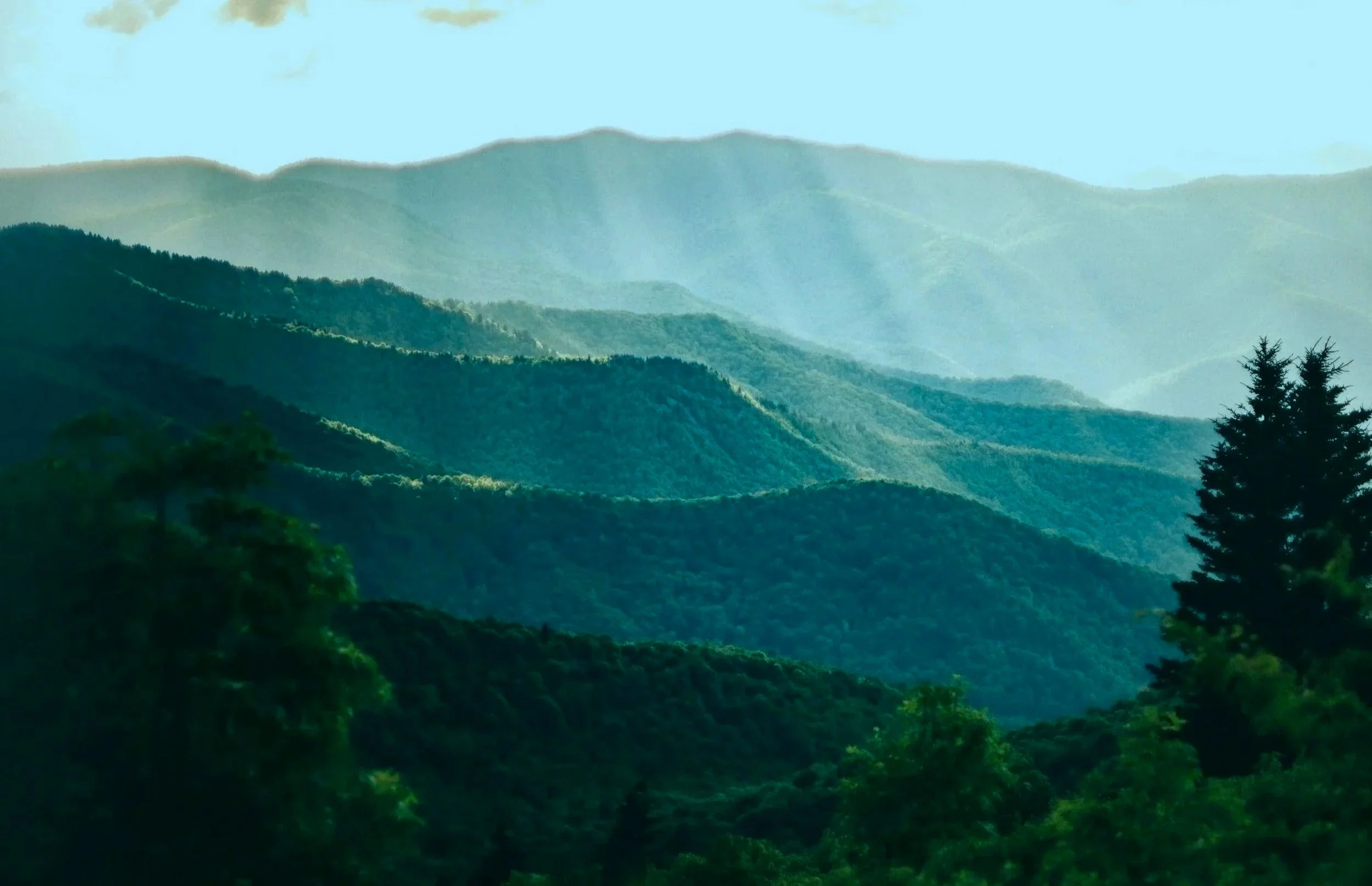 A scenic view of multiple layers of rolling green mountains under a pale blue sky with light clouds.