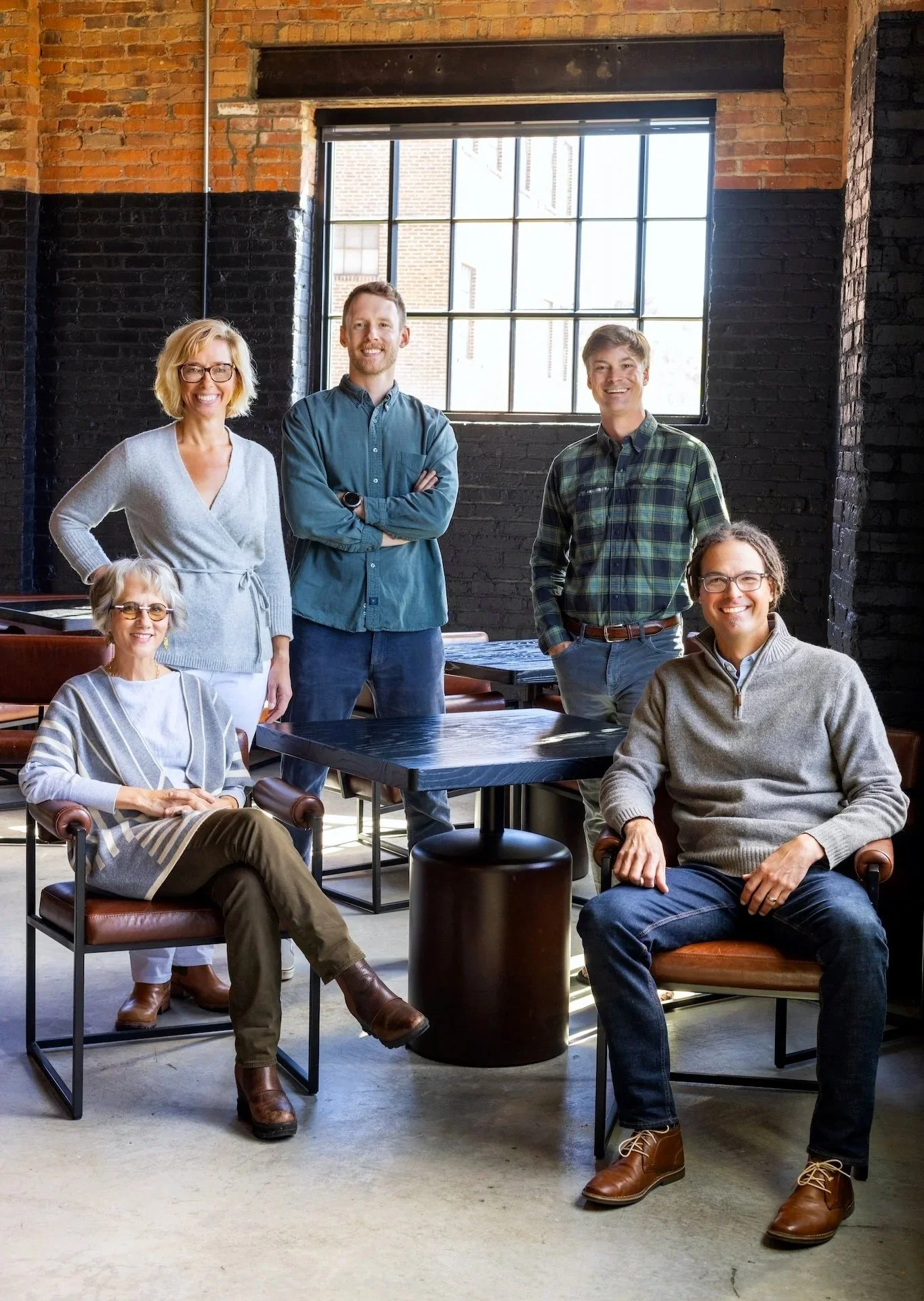 A group of five people, three men and two women, are in a modern industrial-style office with black-painted brick walls and large windows. They are smiling, with some sitting and some standing, dressed casually.