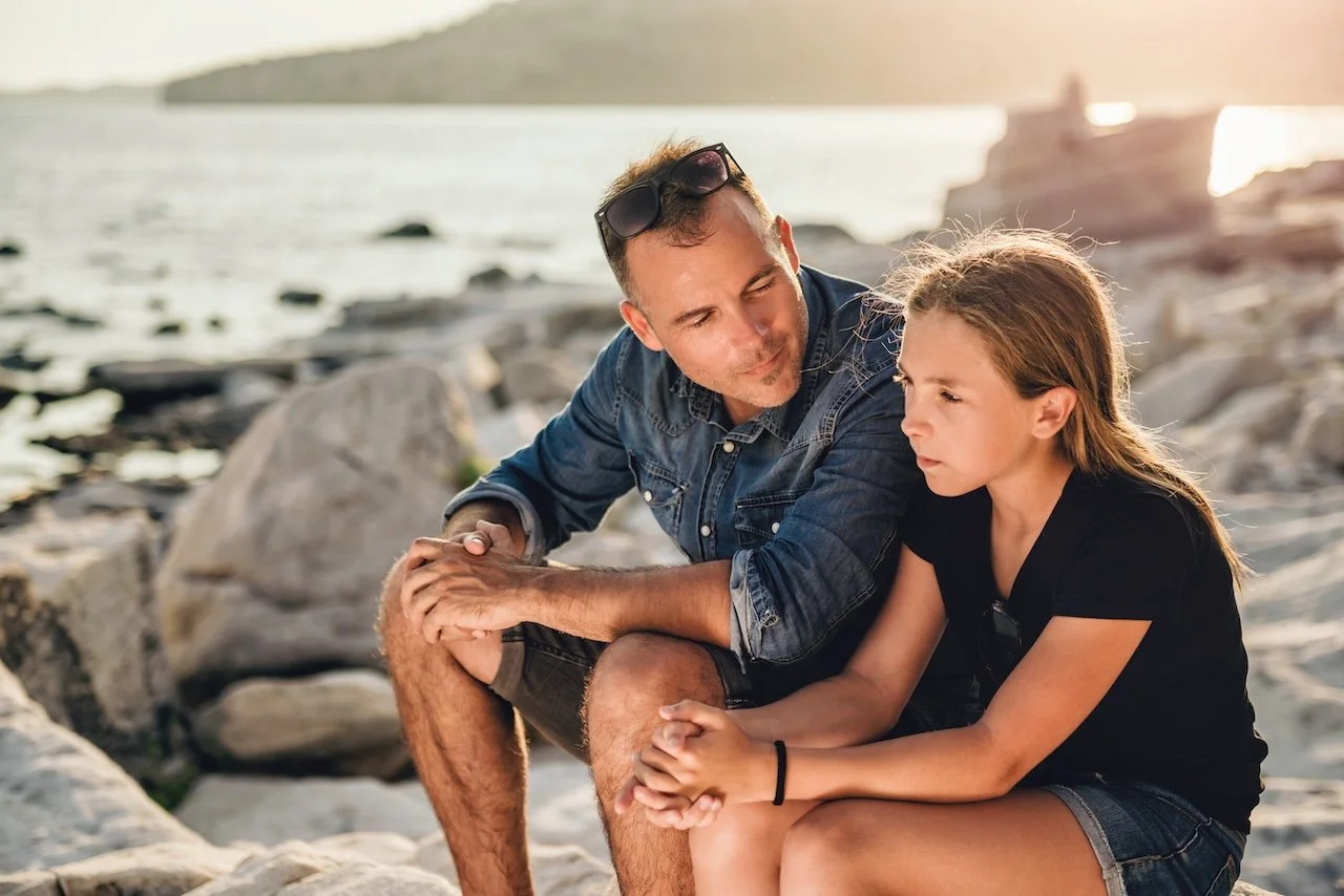 A man and a girl sitting on rocks by the water's edge, having a conversation during sunset.