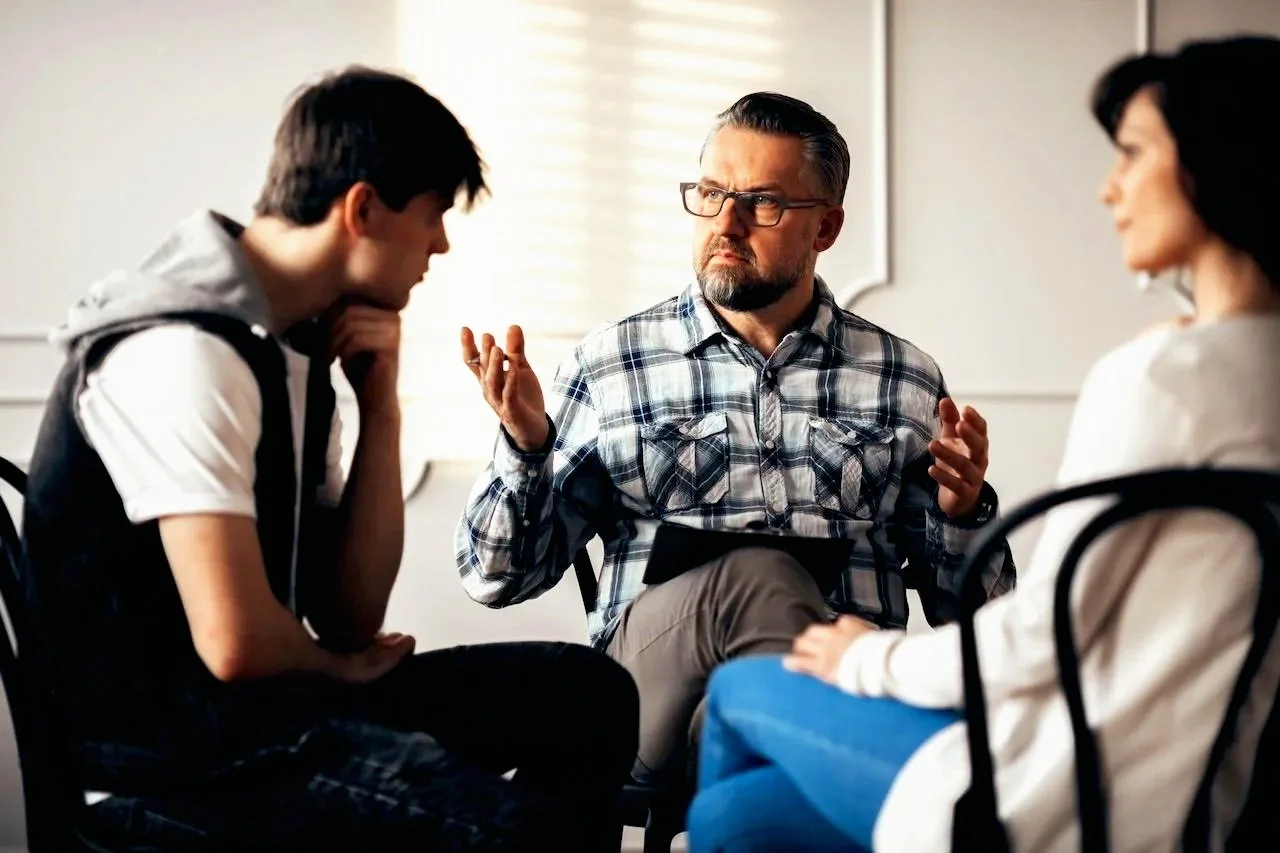A man with glasses and a checked shirt appears to be counseling or talking to a teenage boy and girl, both sitting with attentive expressions in a room with bright natural lighting.