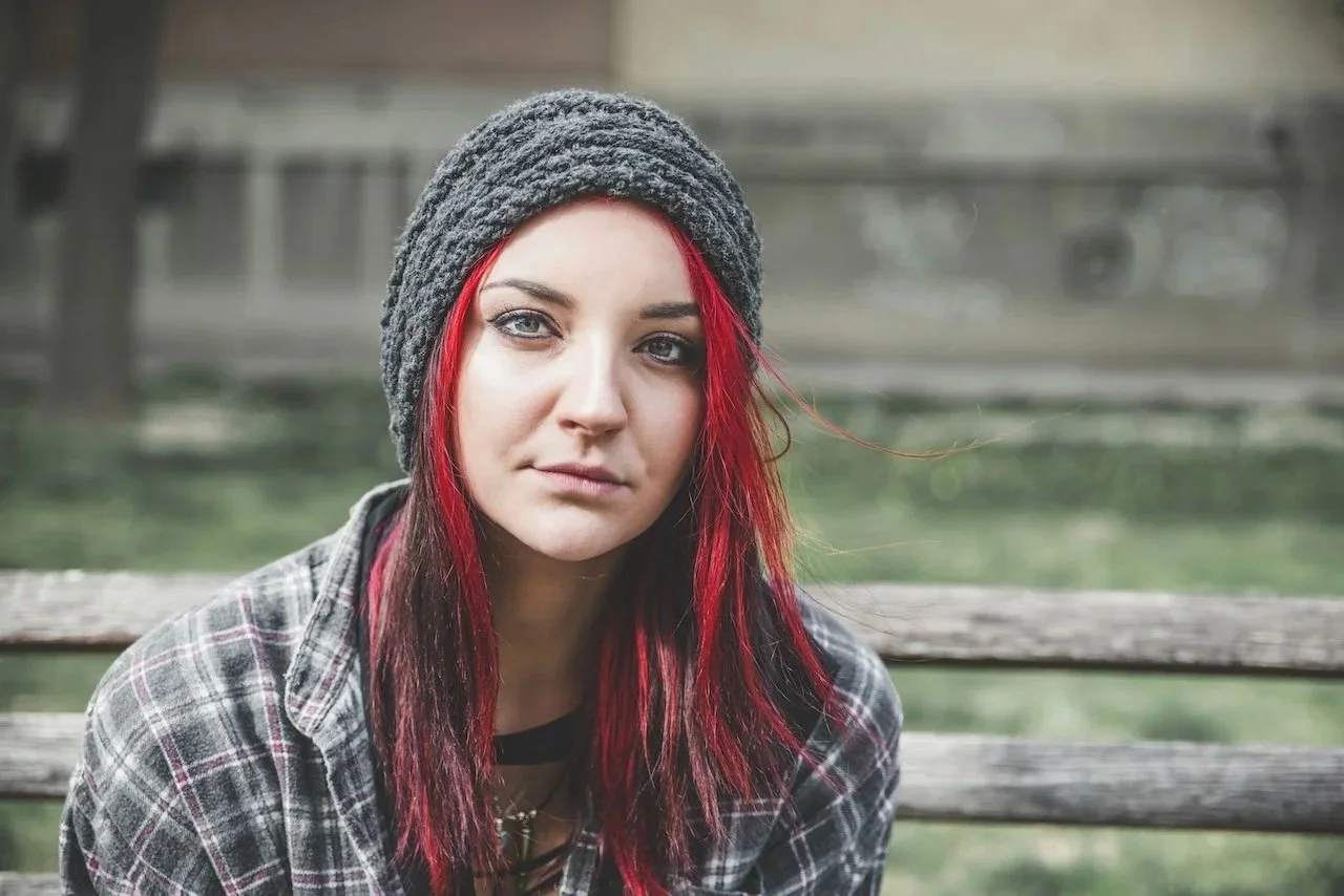Young woman with red and black hair wearing a gray knit hat and plaid jacket, sitting outdoors on a wooden bench with a blurred background.