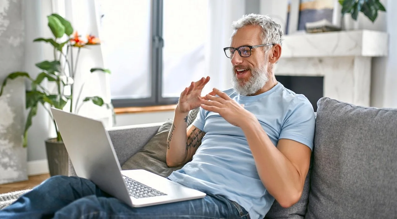 An older man with gray hair, glasses, and a beard sitting on a gray couch with a laptop on his lap in a bright living room. He is smiling and gesturing with his hands. There are green plants and a window with natural light in the background.