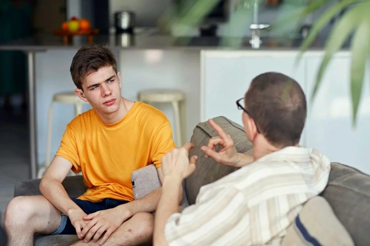 A young man in a yellow shirt sitting on a couch, looking confused or skeptical, as he listens to an older man in glasses and a striped shirt, who is talking and gesturing.
