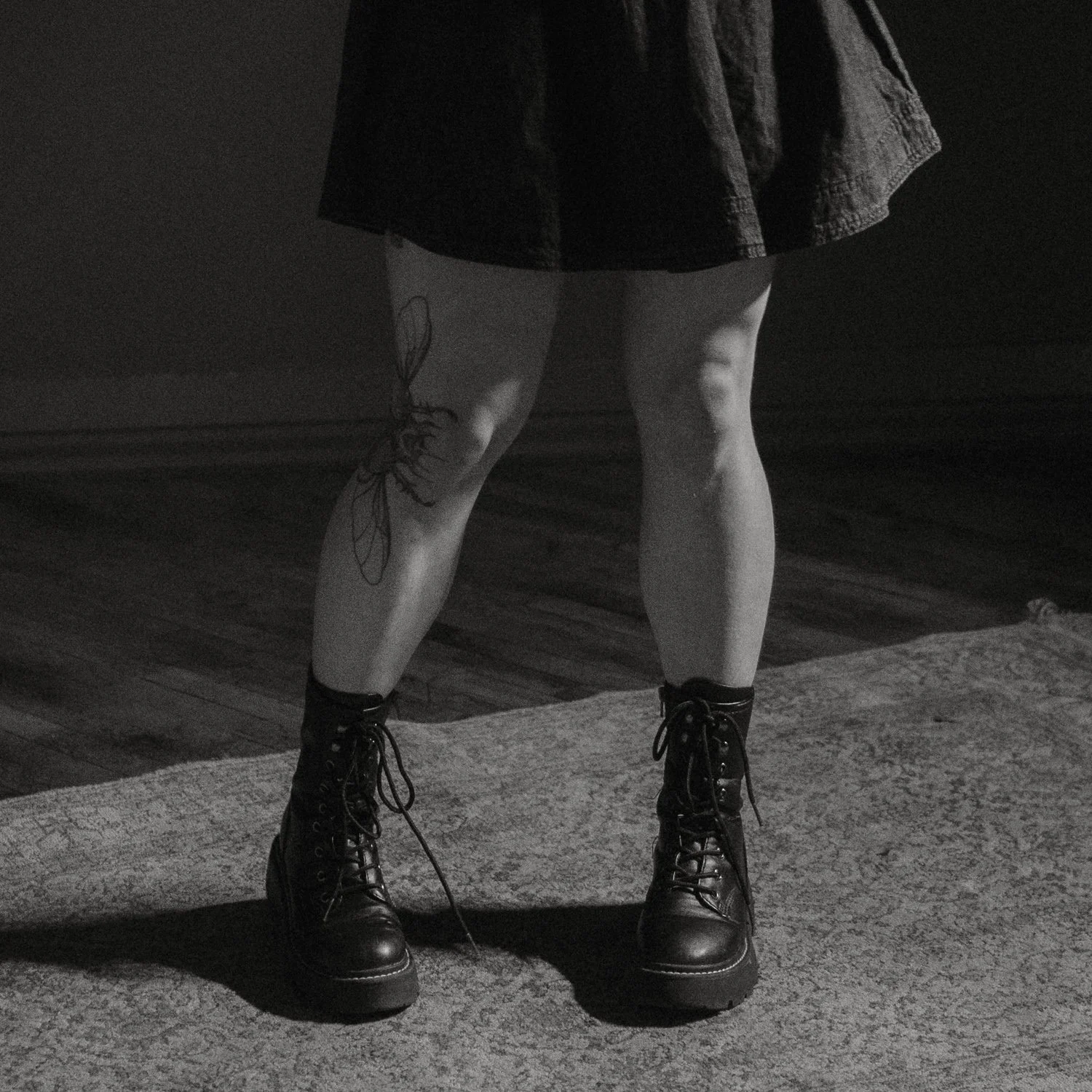 Black and white photo showing a person's legs from the knees down, wearing combat boots and a dark skirt, with a tattoo of a butterfly on one thigh, standing on a rug on a wooden floor.
