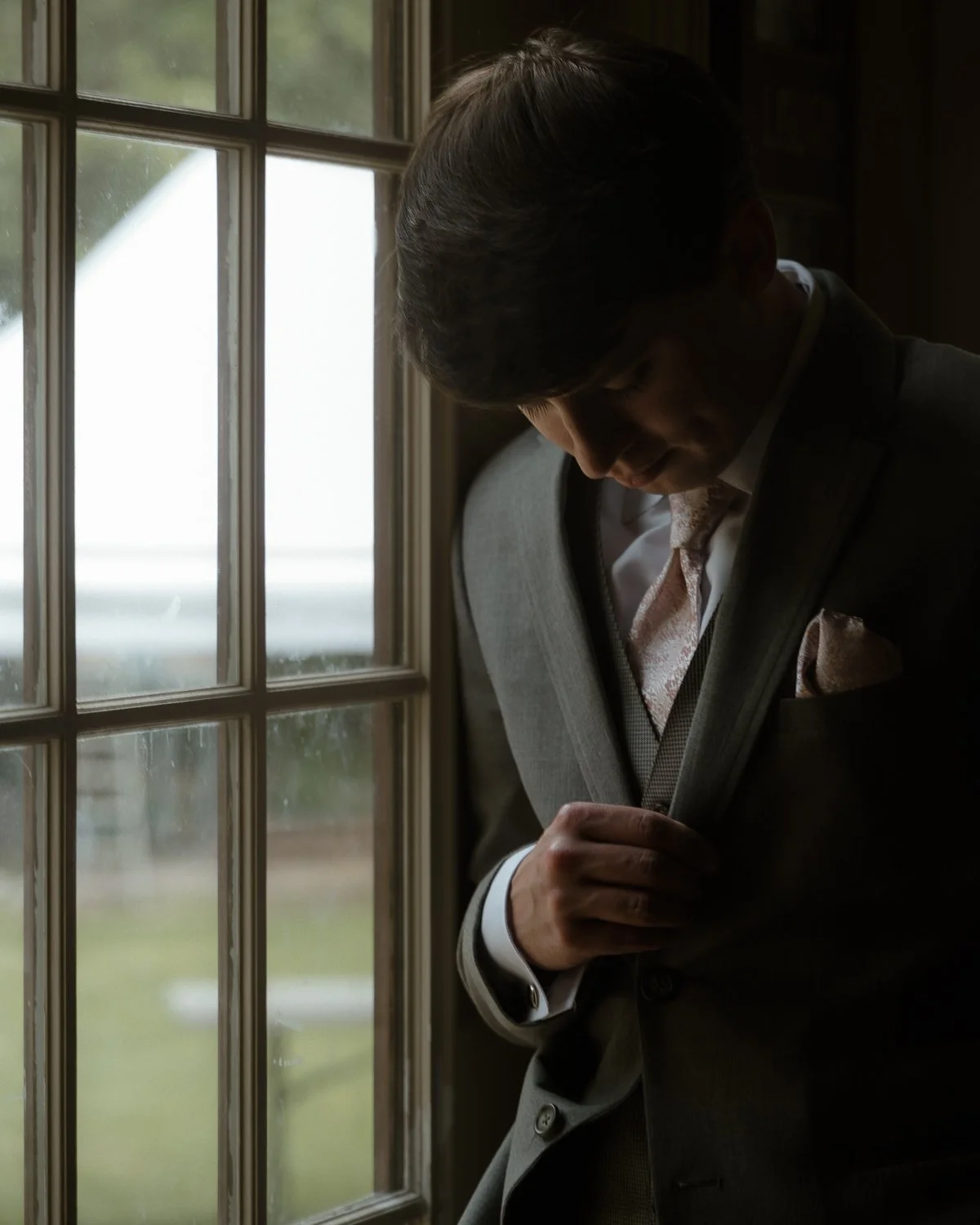 A man adjusting his suit jacket near a window, with a blurred outdoor scene in the background.