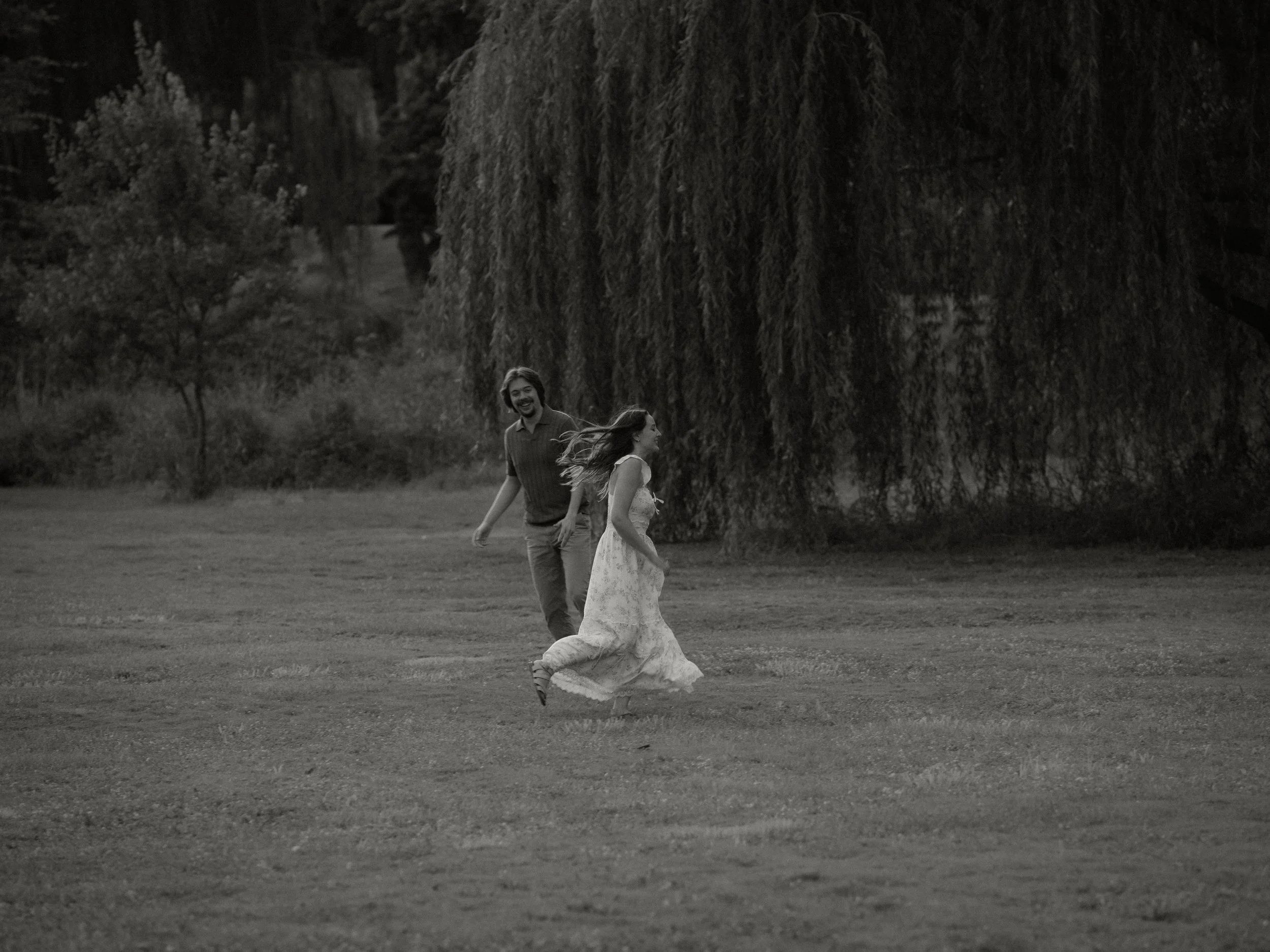 A black-and-white photo of a smiling man and woman walking outdoors on grass near trees, with the woman in a flowing dress and the man in casual attire, in front of large hanging branches of a tree.
