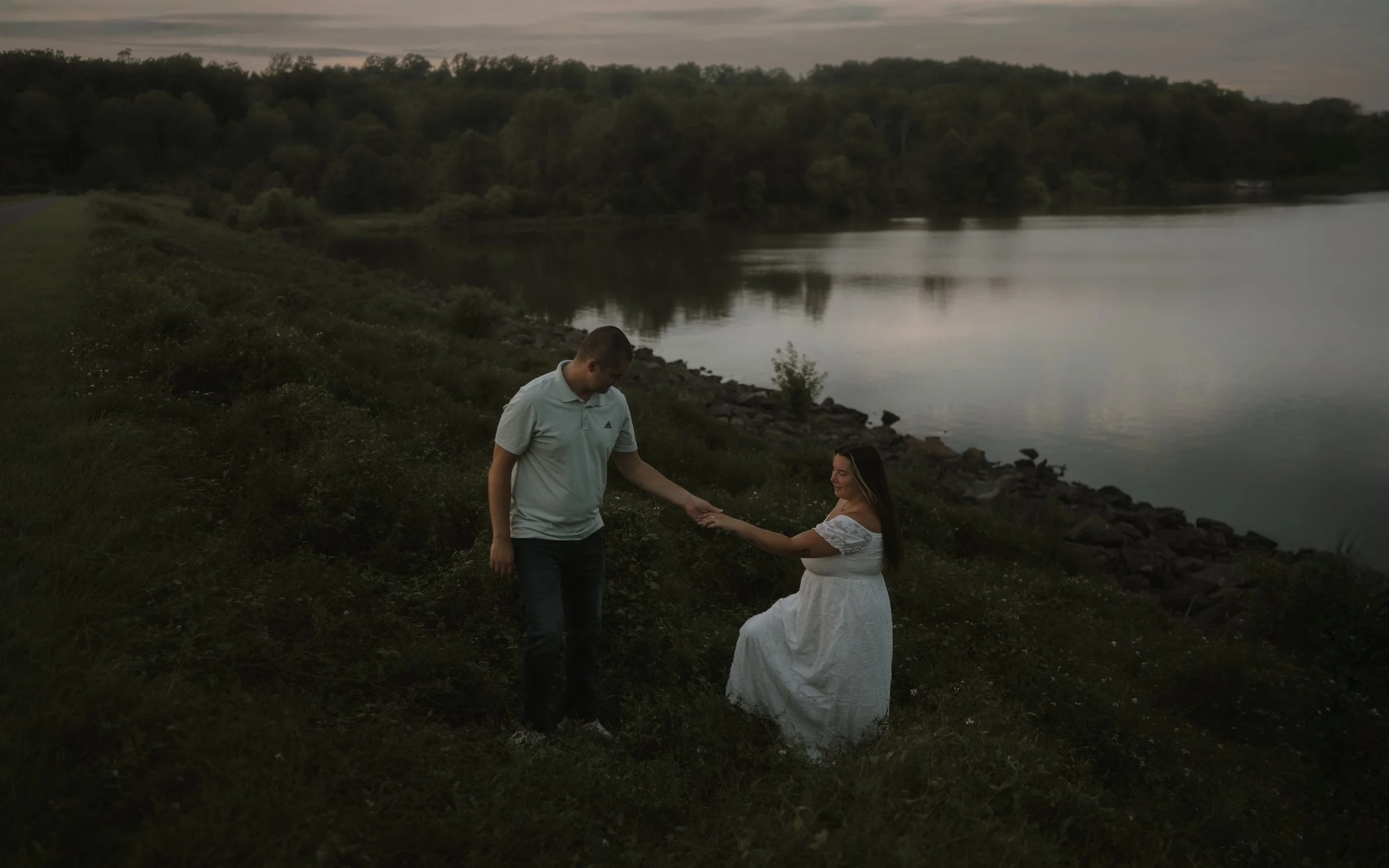 A couple holding hands by a river during dusk, with trees in the background.