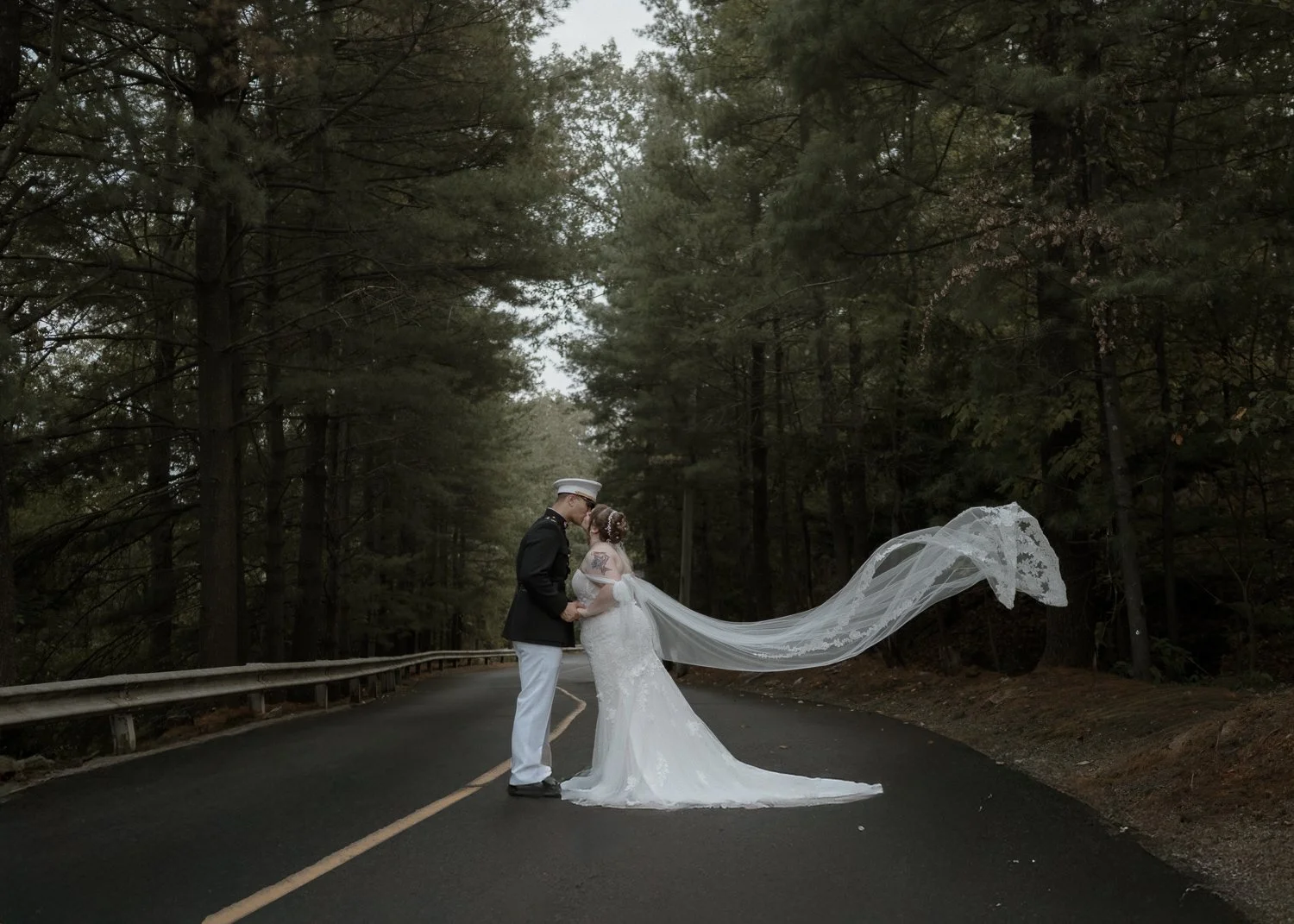 A bride and groom in wedding attire standing on a forest road, holding hands and kissing. The bride's long veil extends behind her, flowing into the air. The scene is surrounded by tall trees on both sides of the road.