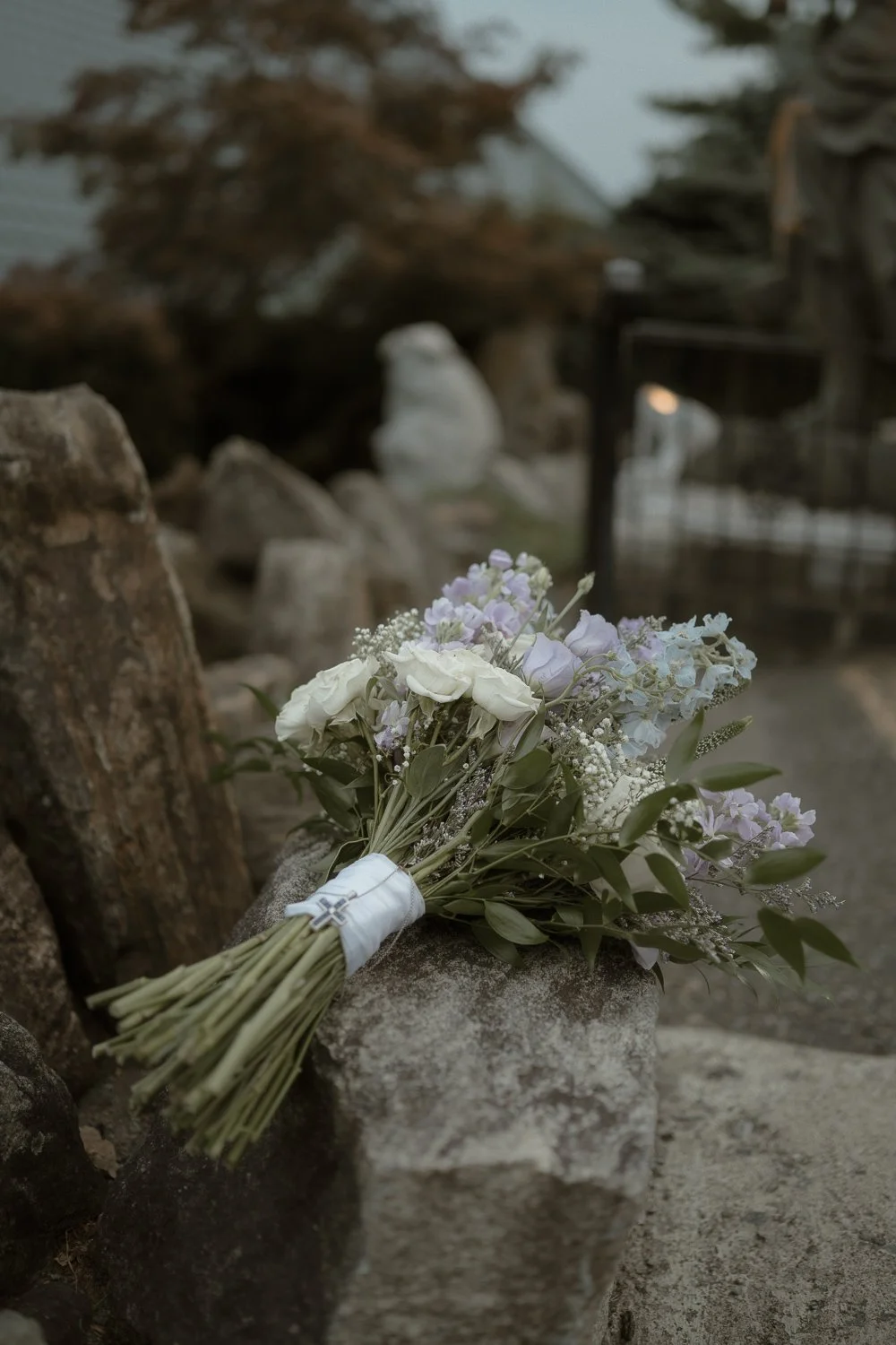 A bouquet of white and pastel purple flowers resting on a stone surface outdoors with rocks and trees in the background.