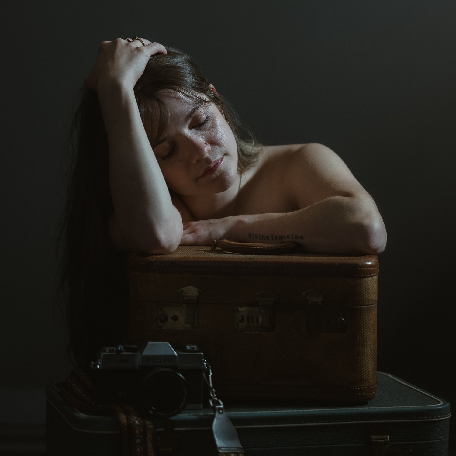 A woman with brown hair and a nose ring resting her head on her hand on top of a vintage suitcase, with a camera in front of her, in a dark setting.