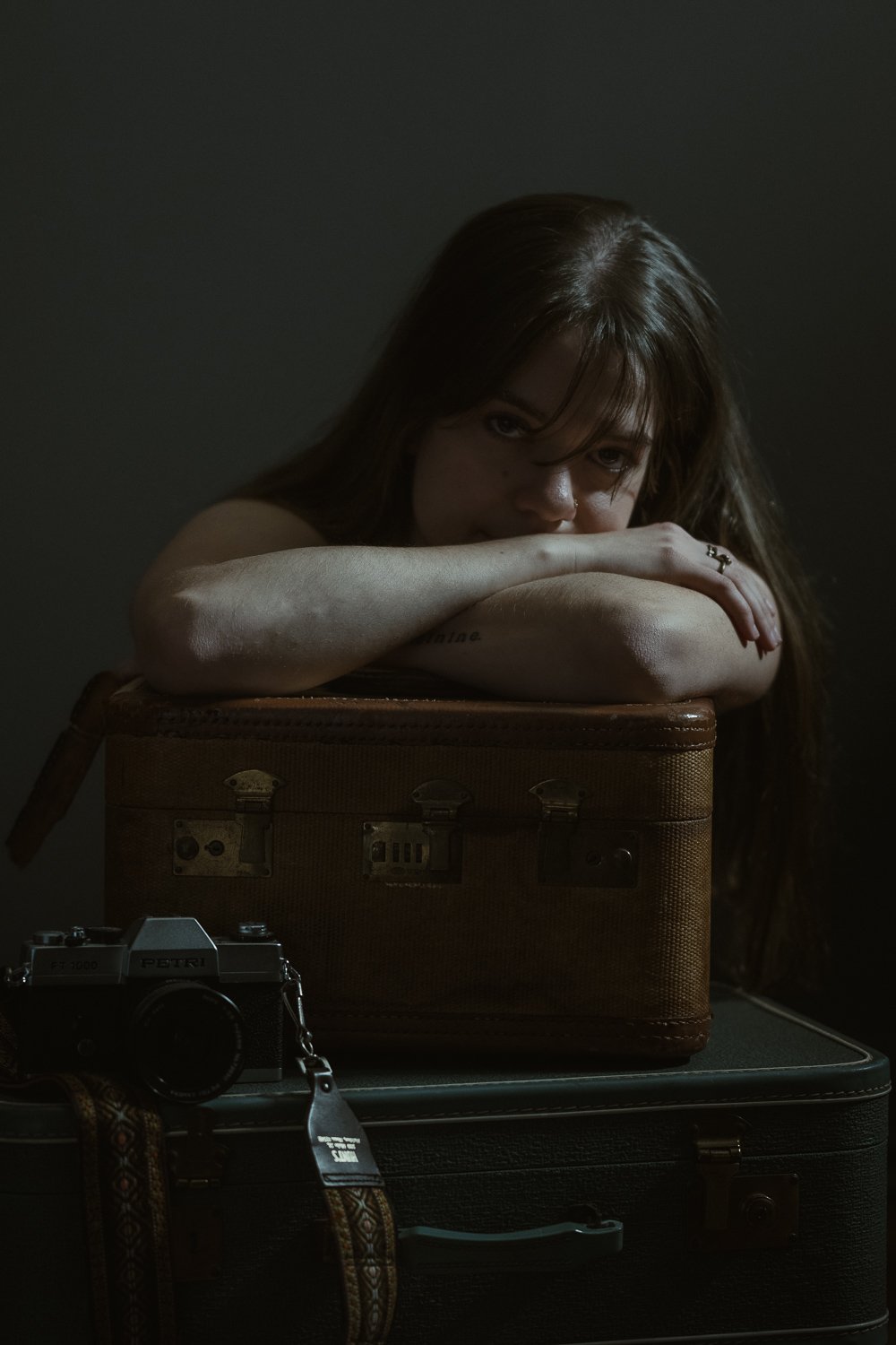 A woman with dark hair resting her head on her arms, leaning on vintage suitcases, with a camera in front and a dark background.
