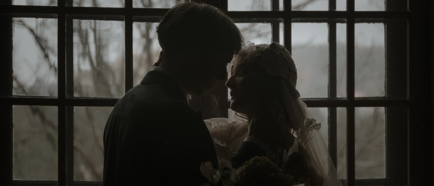 Silhouettes of a man and woman facing each other closely in front of a window, with the woman holding a bouquet and wearing a veil and floral headpiece, suggesting a romantic or wedding scene.