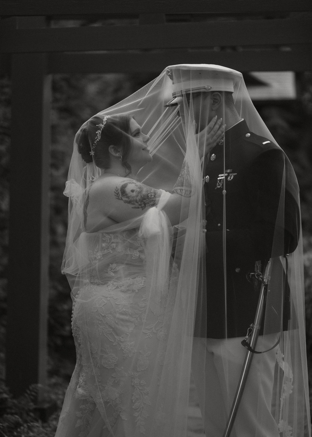A bride wearing a wedding gown and veil, embracing a groom in a military uniform under a canopy, with her hand on his face, intimacy and love shown in the moment.