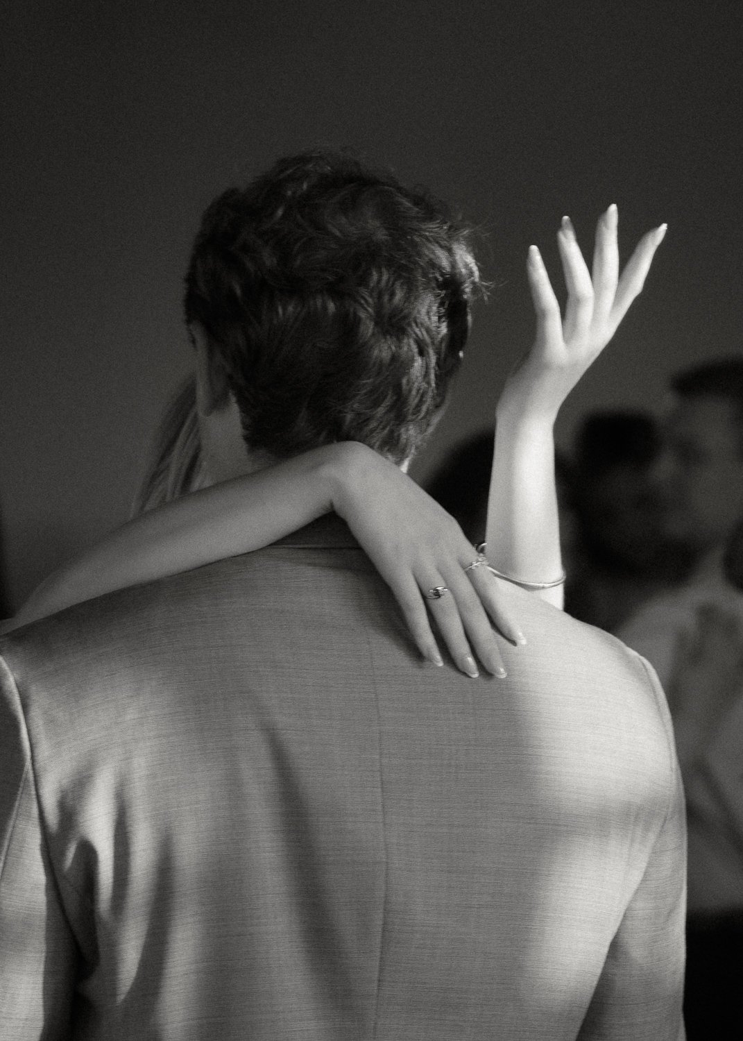 A black and white photo of a man in a suit with a woman embracing him from behind, her arm around his shoulder, and her hand raised in the air, with a woman in the background slightly out of focus.