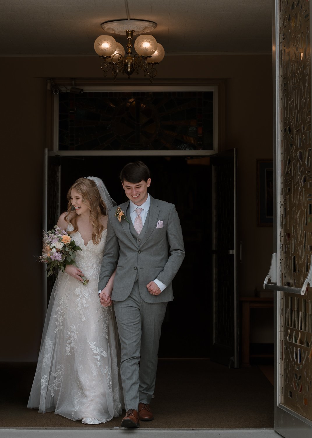 A bride and groom walking hand in hand out of a building, smiling happily. The bride is wearing a white lace wedding dress and holding a bouquet of flowers, and the groom is in a gray suit with a pink tie and boutonniere.