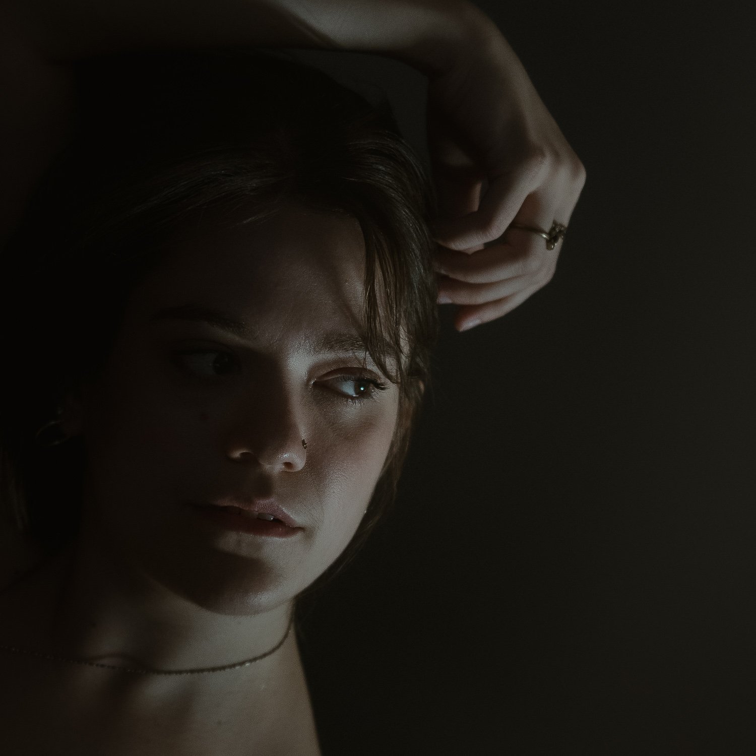 Close-up of a woman with short hair and a nose piercing, gazing to her left, with her hand resting on her forehead, against a dark background.