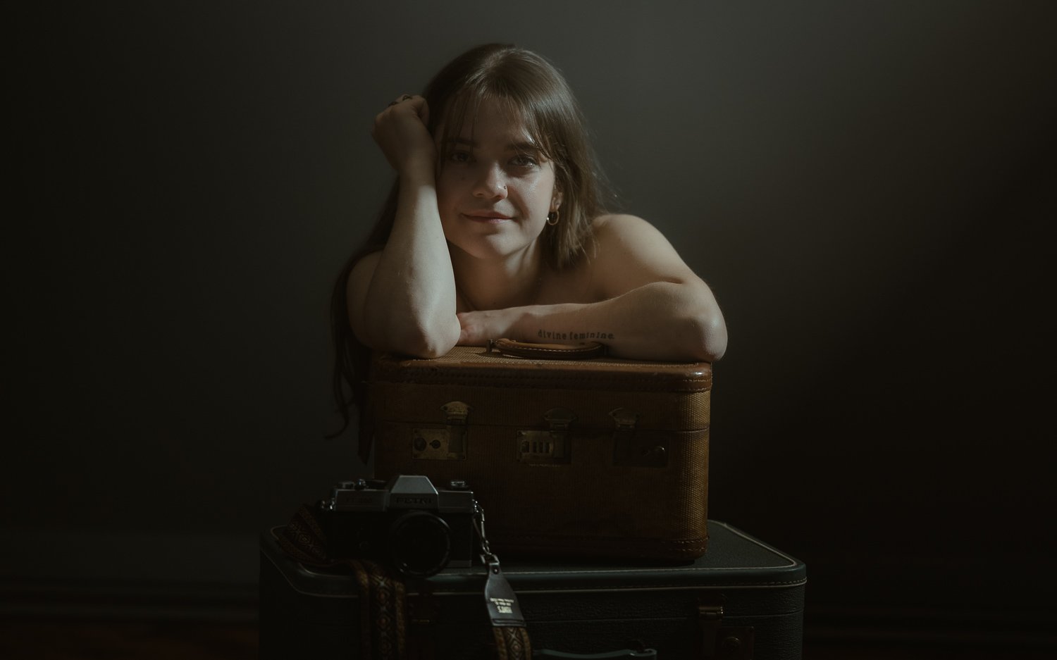 A woman with shoulder-length hair rests her head on her arm, leaning on a vintage suitcase. There is a camera in front of the suitcase on a surface, set against a dark background.