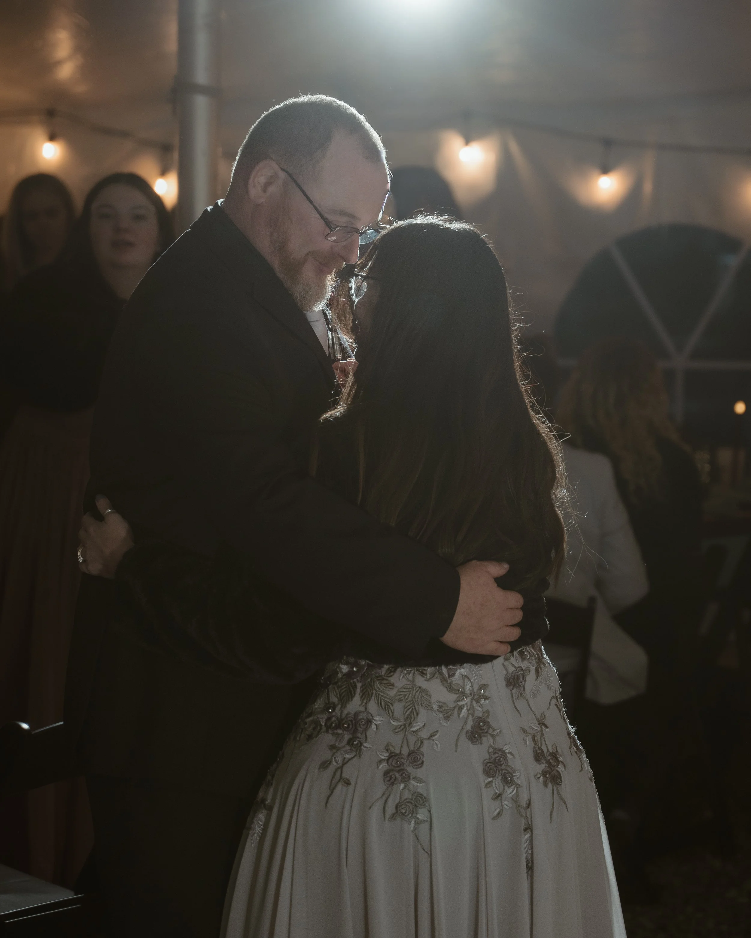 A man and woman embrace while dancing at a dimly lit event, with other guests visible in the background.