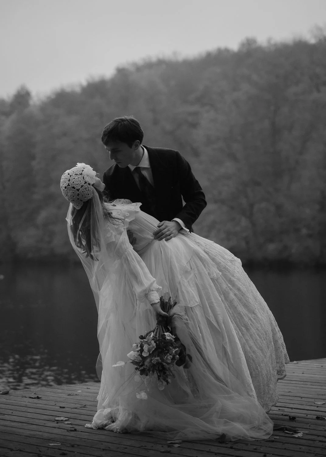 A black and white photograph of a bride in a wedding dress holding a bouquet, being dipped backward by a groom in a suit on a wooden dock near a body of water with trees in the background.