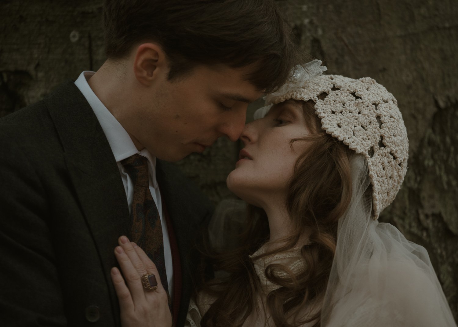Close-up of a man and woman with noses touching, eyes closed, against a rustic wooden background. The man is dressed in a dark suit and white shirt, and the woman is wearing vintage-style clothing, including a lace hat and a ring with a large stone. The scene is intimate and emotional.