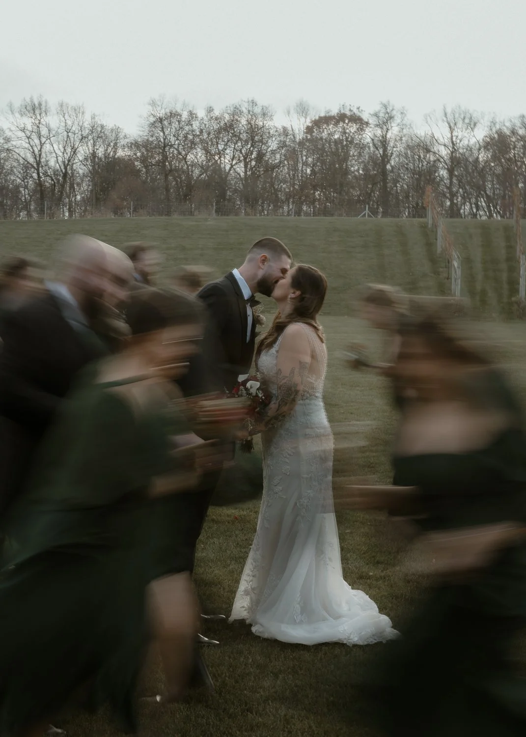 A bride and groom are kissing during their wedding ceremony outdoors, with blurred guests around them, on a grassy field with trees in the background.