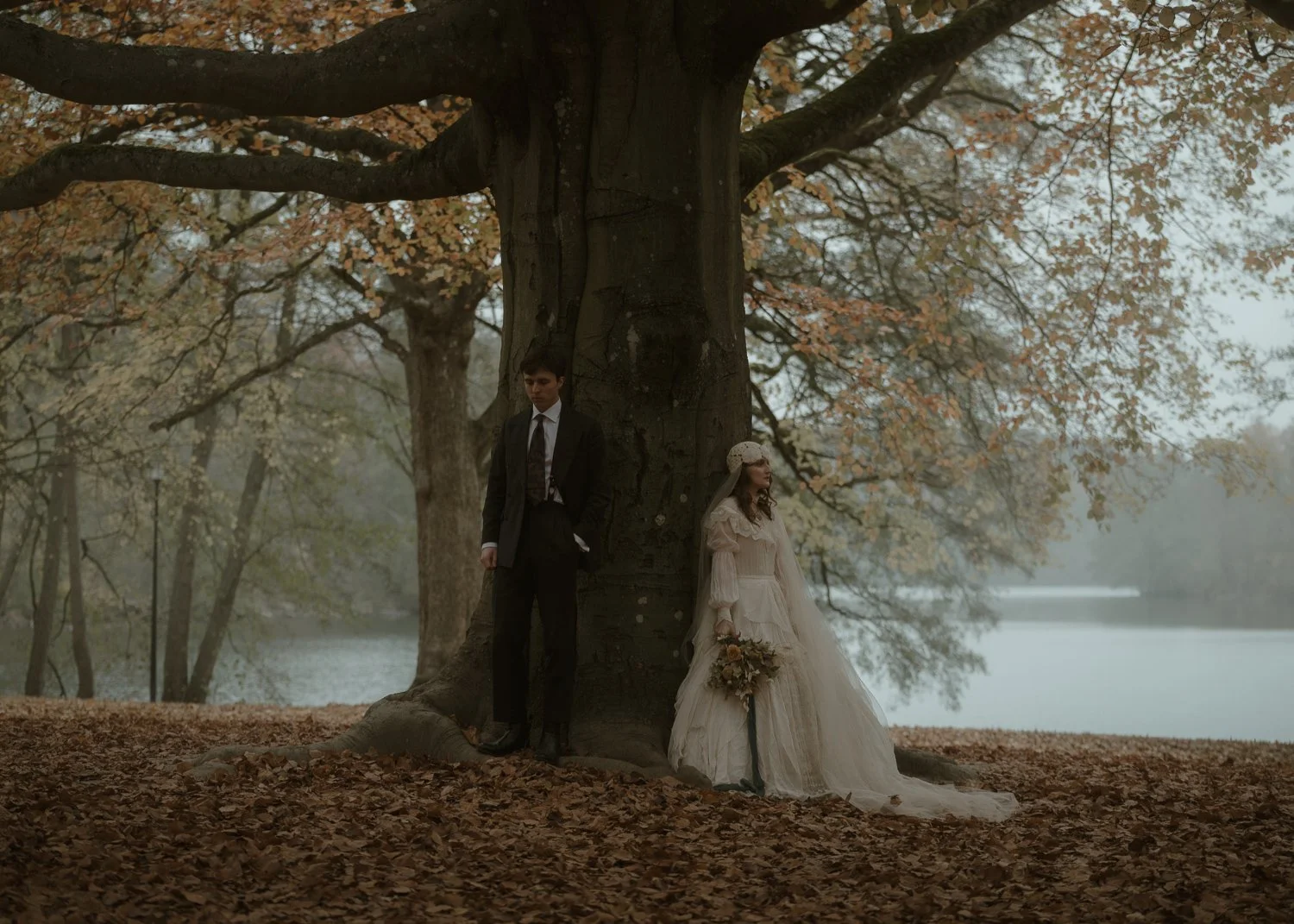 A man and woman stand on opposite sides of a large tree in an autumnal park, with the woman holding a bouquet of flowers. The woman is wearing a vintage-style white dress and a headpiece, while the man is dressed in a dark suit and tie. The scene is foggy with fall leaves on the ground and trees in the background.