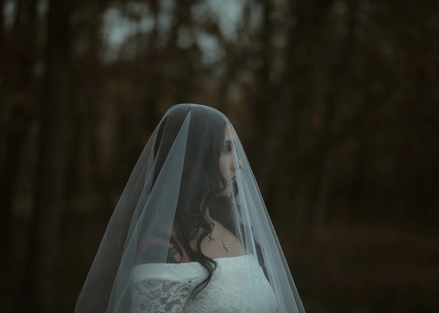 A woman in a white wedding dress with lace details stands outdoors, her face turned sideways, covered by a sheer veil, with a dark, blurry forest background.